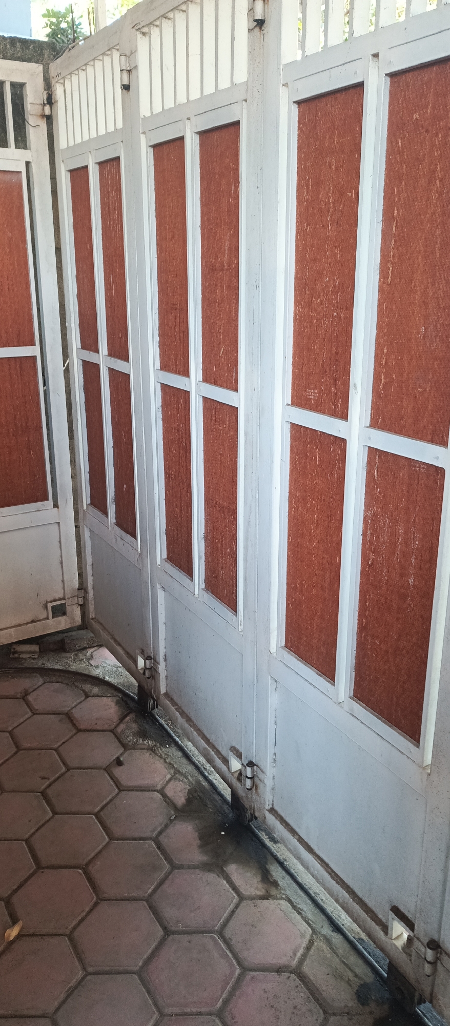 This image shows a close-up of a weathered, white metal gate with reddish-brown panels. The gate appears to be composed of multiple sections, each with vertical dividers. The panels have a textured surface and are slightly faded. The gate is positioned outdoors, with a glimpse of greenery at the top left. The ground in front of the gate is paved with hexagonal tiles, and there is some wetness and debris on the ground near the base of the gate. The scene suggests an outdoor area, possibly a pathway or entrance. The location in Arjasa, Indonesia, is context provided and not visually discernible from the image itself.