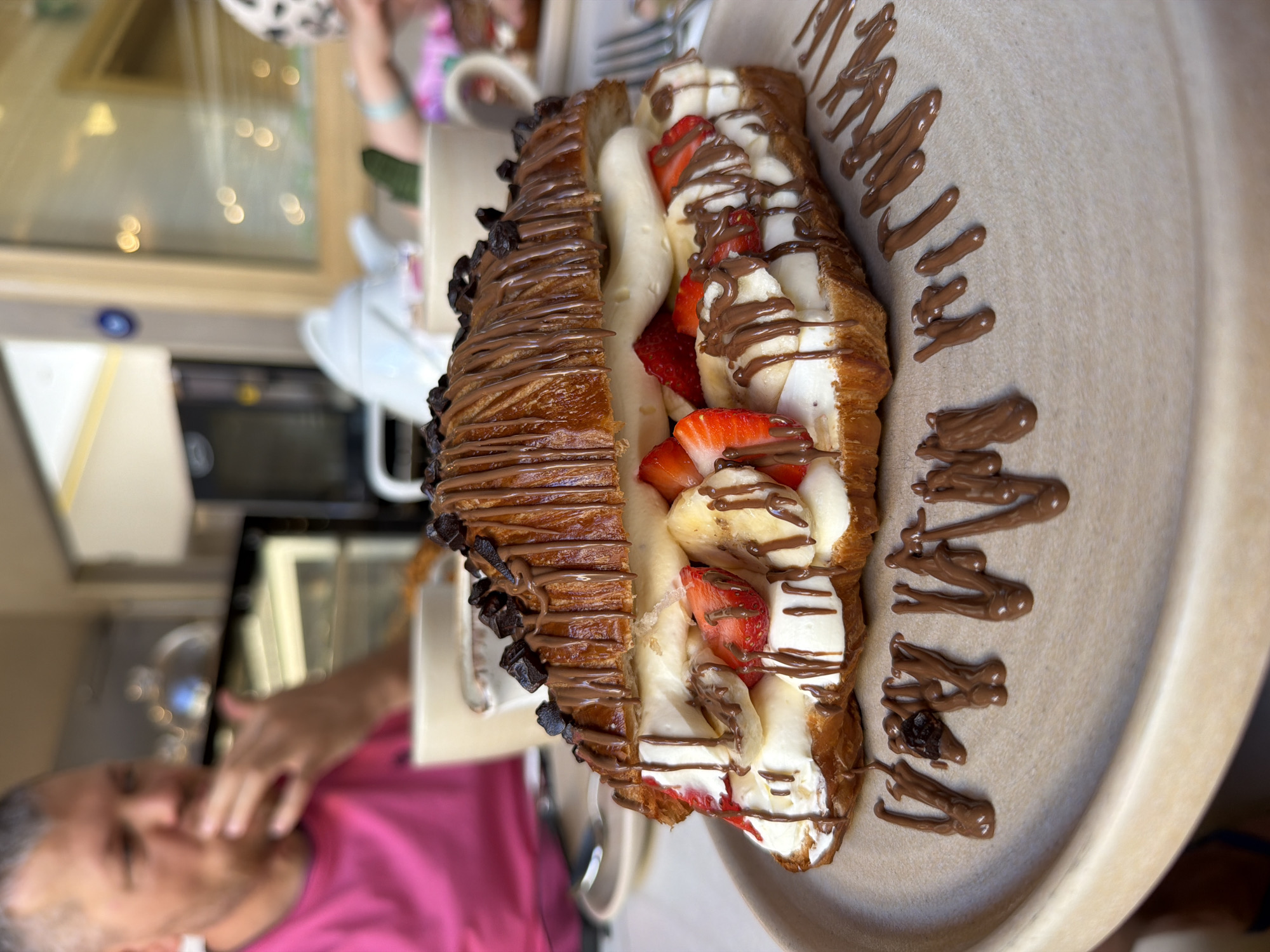 This image captures a close-up of a lavishly prepared dessert or breakfast item, set in an indoor cafe environment in Fethiye, Turkey. The primary subject is a large, elongated croissant, presented split open and filled, resting on a light beige, textured platter.
The croissant, golden-brown and flaky, is generously stuffed with sliced fresh strawberries and bananas, interspersed with a creamy white filling, likely whipped cream or a sweet spread. Both the exterior and the filling are drizzled with dark brown chocolate sauce, enhancing its visual appeal. Along the left edge of the croissant, additional dark chocolate chips or chunks are visible.
To the right of the croissant, artfully written in chocolate sauce directly on the platter, is the word "MAMACITA".
The background is softly blurred, emphasizing the food in the foreground. In the bottom left, a person wearing a bright pink shirt is visible, their hand near their face, indicating they are part of the dining scene. Another person, partially visible and blurred, can be seen in the top right background, wearing a white and black spotted head covering. A white ceramic cup with a handle is also discernible in the mid-background. Blurry reflections in what appears to be a window or display case are also visible in the upper background.
The overall scene suggests a pleasant, casual setting, likely a cafe or restaurant, where people are enjoying a sweet indulgence. The lighting is bright, indicative of daytime.
