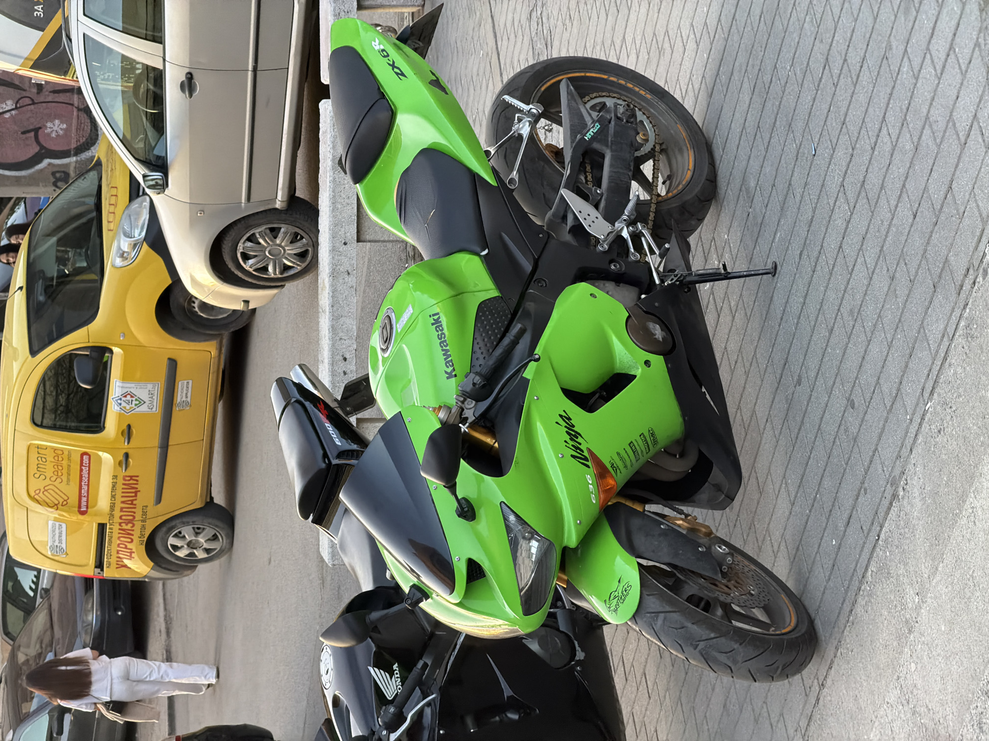The image shows a street scene in Sofia, Bulgaria, featuring several vehicles parked alongside a sidewalk. Prominently displayed is a bright green Kawasaki Ninja 636 motorcycle, parked perpendicular to the curb. Behind it, partially obscured, is a black Honda motorcycle.
To the left of the motorcycles, a yellow van is parked, displaying the logo of "Smart Sealed International London" and related Bulgarian text about waterproofing ("УИДРОИЗОЛАЦИЯ"). Adjacent to the yellow van is a beige car, and further to the left, a partially visible black car is present.
In the lower-left corner, a person dressed in white stands beside a dark-colored car, facing away from the camera. Graffiti is visible on a wall or building in the background, behind the cars. The street is paved with interlocking concrete blocks.