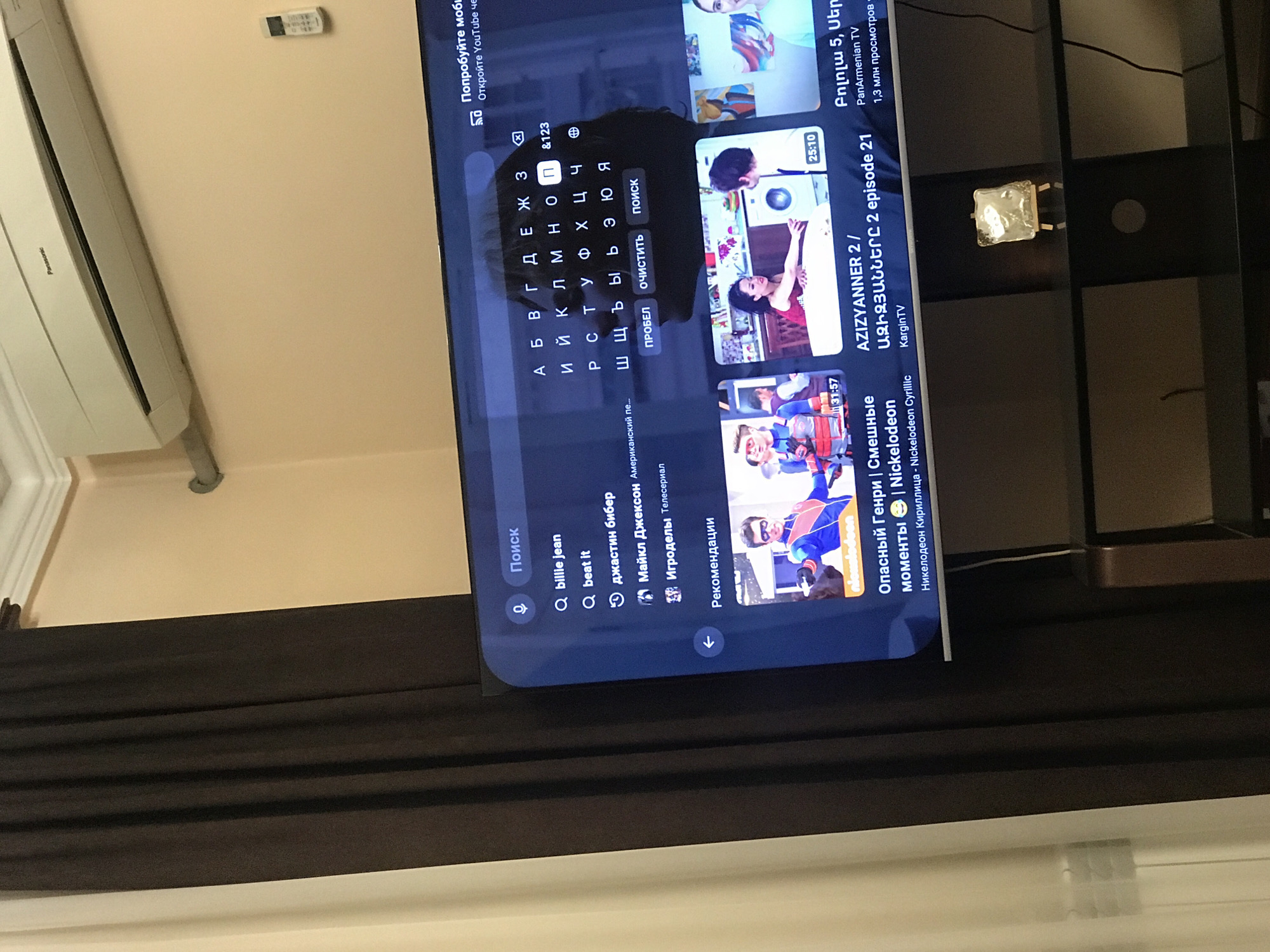 The image shows a television displaying a search interface on YouTube in what appears to be a residential setting. The search query is set to "billie jean". In addition to the search bar, there are recommendations for "beat it" and Justin Bieber. The search keyboard is in Cyrillic. On the screen, some of the recommended content includes "AZIZYANNER 2 /...", "Dangerous Henry". A remote control is visible in the top right. There is an air conditioning unit on the wall. The location is likely Yerevan, Armenia given the Cyrillic letters.