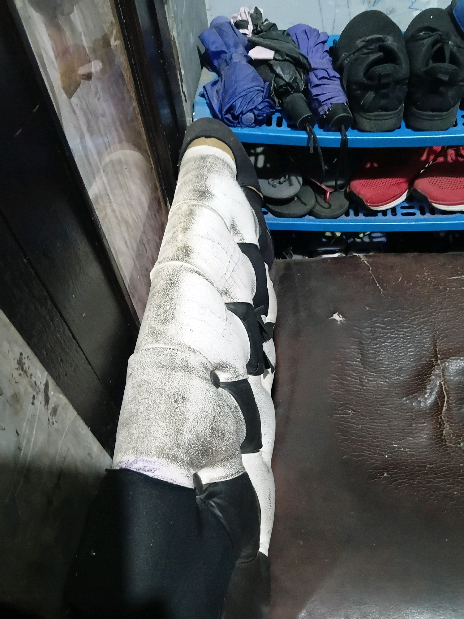 The image shows a close-up of a person's bandaged leg, which is resting on a dark brown, possibly leather, surface. The leg is heavily wrapped in white bandages, with some areas appearing dirty or stained. Black fabric is visible at the top of the bandage and extending downwards. In the background, a blue plastic shoe rack is visible, holding several pairs of shoes and folded umbrellas. The shoes appear to be athletic shoes and sandals. To the left of the shoe rack, a dark, reflective surface, possibly a mirror or glass, shows a partial reflection of the environment, including what appears to be another person. The overall environment appears to be indoors, likely a room in a home or establishment in Cikal, Indonesia. The scene is mundane and does not depict any specific event or interaction.