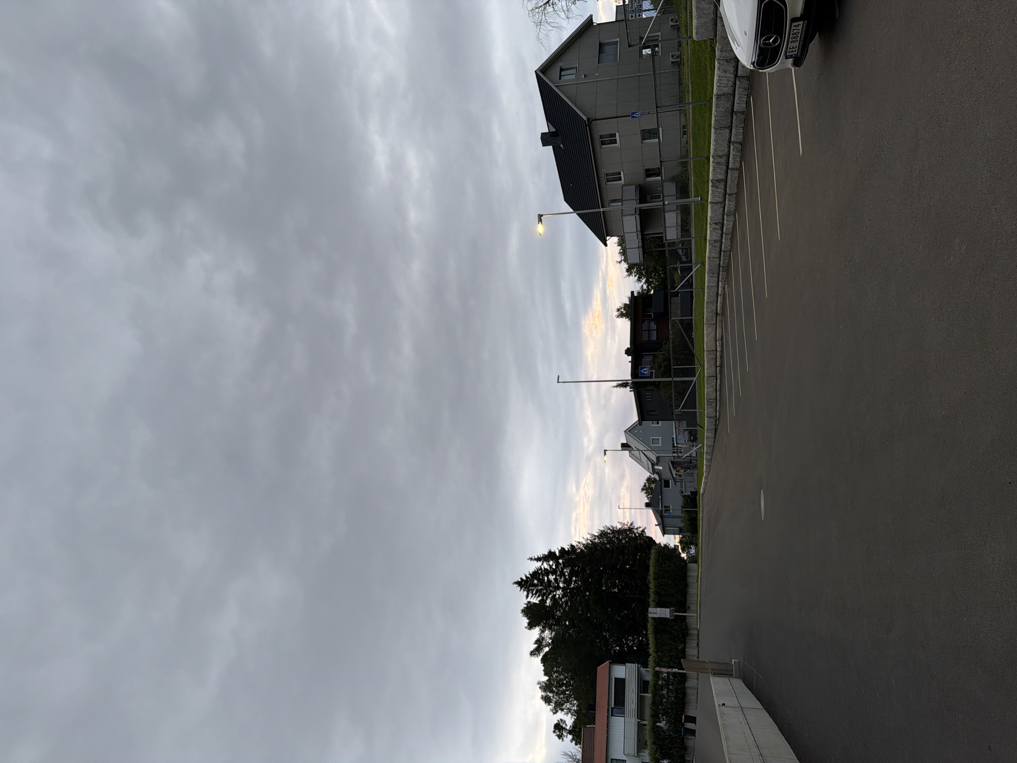 This image depicts an outdoor scene in the city of Harstad, Norway, likely during twilight. The foreground features a dark asphalt parking lot with white parking space markings. A white Mercedes-Benz car is partially visible on the right side of the parking lot, with its license plate reading "EE 88874".
Beyond the parking lot, a low stone wall separates it from a grassy area with a fence. Behind the fence, several houses are visible, characteristic of a residential neighborhood. These houses are of varying architectural styles and colors, with some having dark roofs and grey or white facades. Trees, including tall, dark evergreen trees, are interspersed between the houses and along the property lines. Streetlights are present, with one illuminated lamp post in the middle ground.
The sky is overcast with heavy, grey clouds, suggesting a potentially cool or damp evening. The overall atmosphere is subdued, typical of a cloudy day or evening in a Nordic region. The image appears to be taken from a slightly elevated position looking towards the houses and the sky. There are no visible people or ongoing activities in the scene.