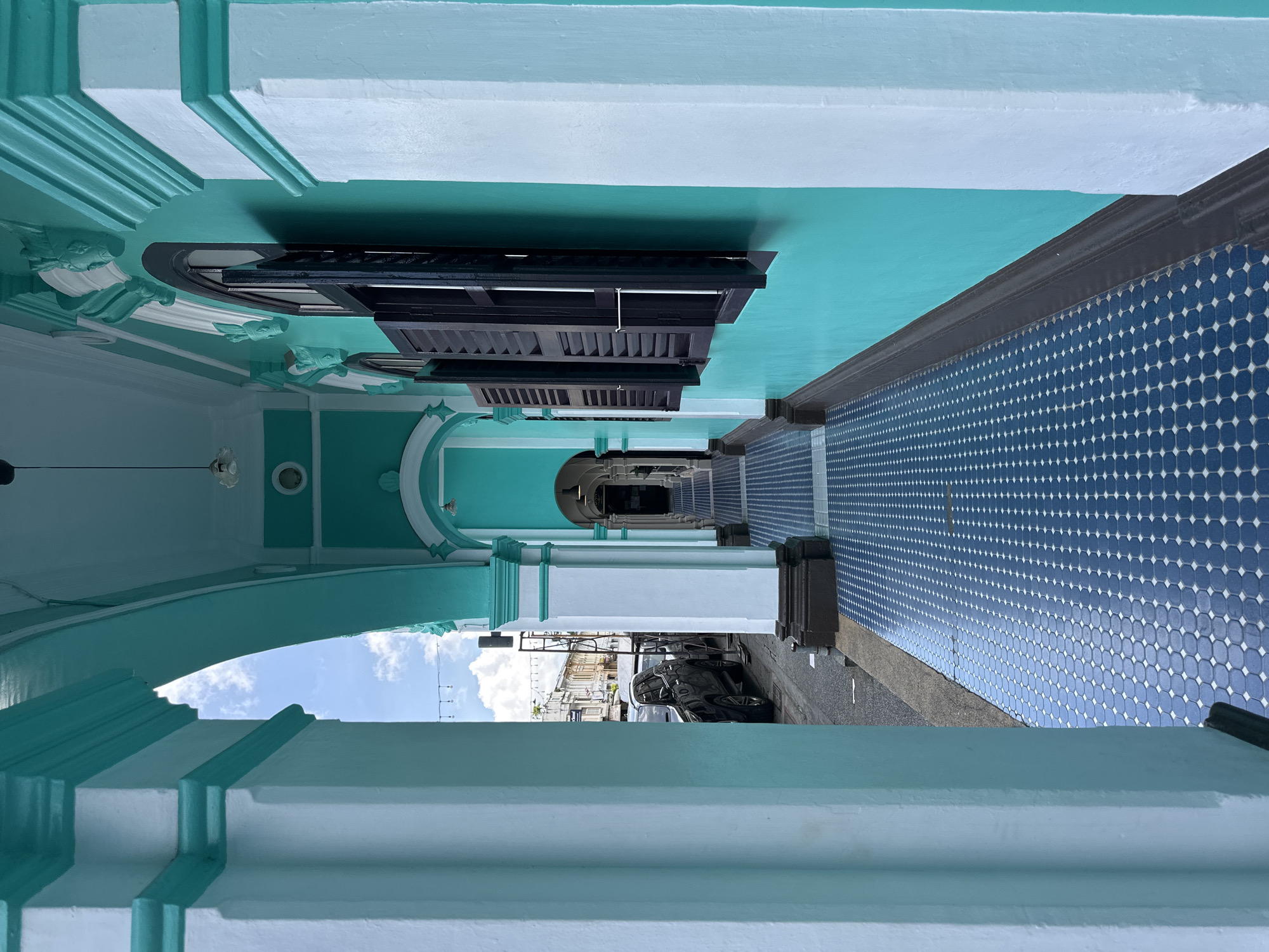 The image depicts a turquoise-colored colonial-style building with white pillars and arches. The walkway in front of the building is paved with blue and white patterned tiles. The walkway leads to a street where a black car is parked. In the background, there are other buildings and some overhead wires. The sky is blue with white clouds. The architecture suggests a historical area, likely within Phuket City Municipality, Thailand, known for its well-preserved Sino-Portuguese shophouses.
