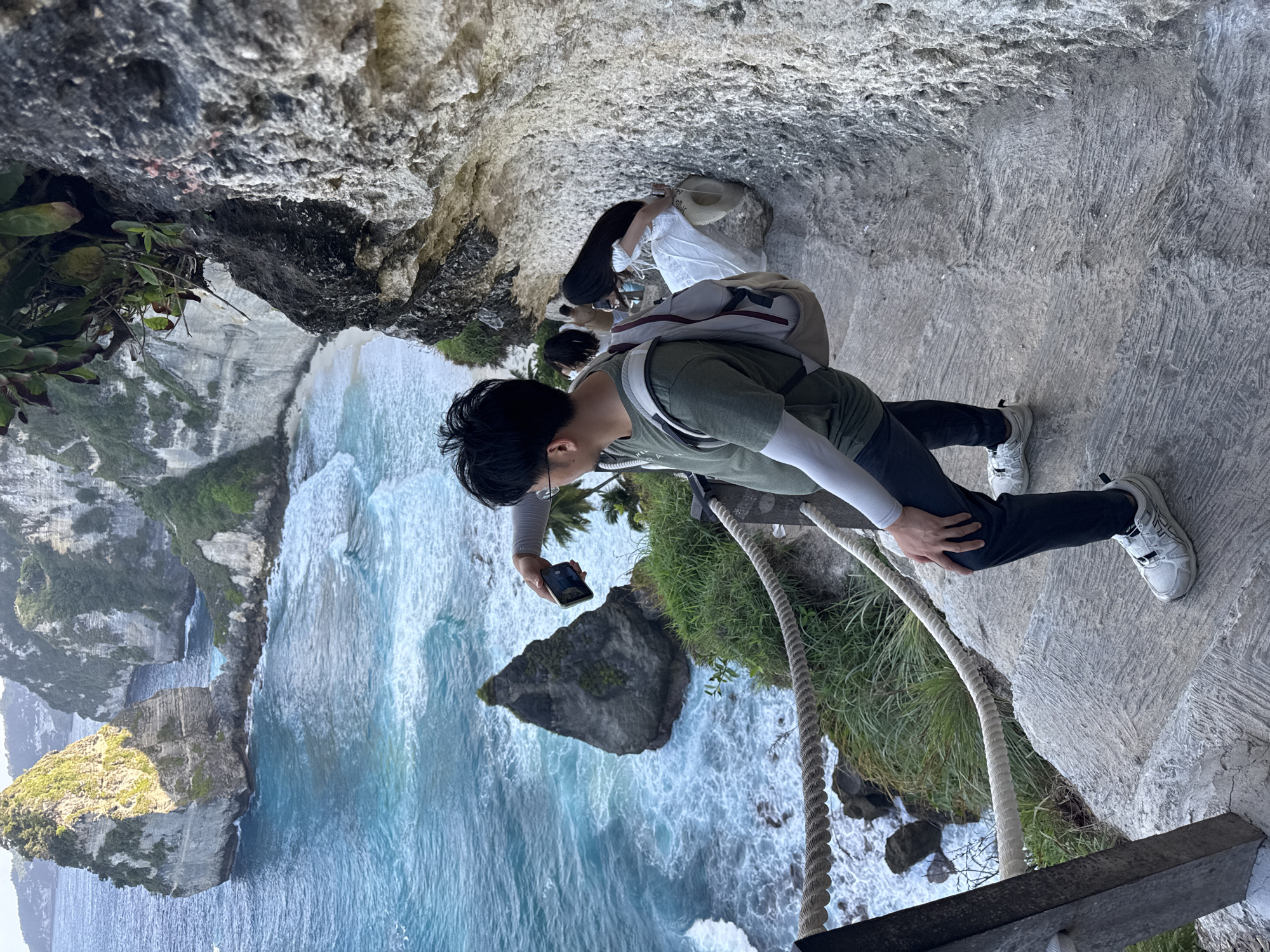 A man wearing a green t-shirt, dark pants, and white sneakers is taking a photo of a rock formation in the ocean with his phone. He has a backpack on and is standing on a stone staircase on a cliffside. Behind him, a woman in a white dress is sitting on the stairs. The ocean is a vibrant blue with waves crashing against the shore. The cliffside is rocky with lush green vegetation. The scene is located at Diamond Beach, Nusa Penida, Indonesia, a popular tourist destination known for its stunning coastal scenery.