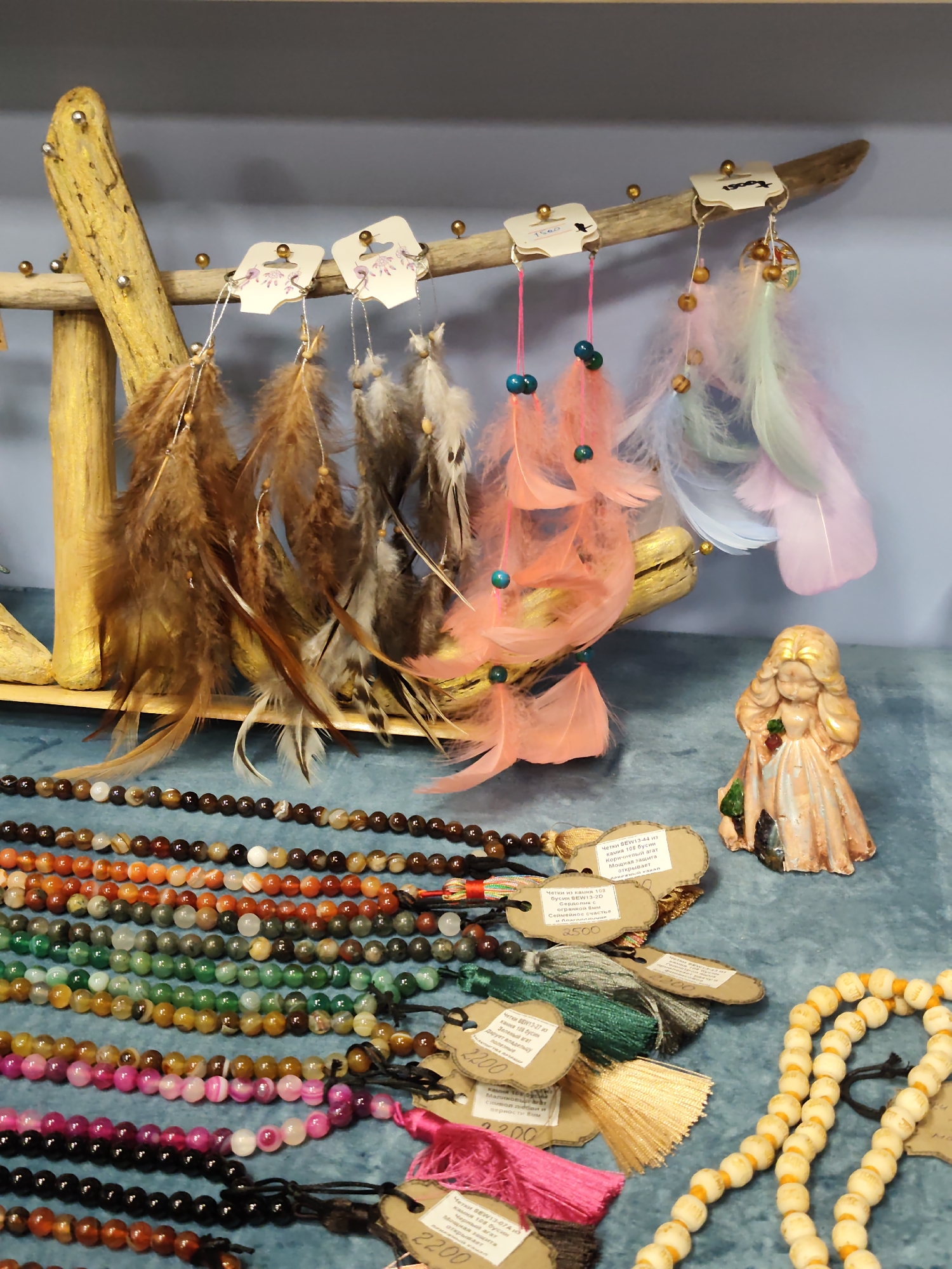 This is a medium shot, eye-level, interior photograph of a display of bohemian-style handicrafts at a street market stall. The stall is located in Sochi, Russia.
In the upper portion of the image, a gnarled, light-colored wooden branch is suspended horizontally, supported by two vertical wooden poles on the left. Several small, brass-colored pins are driven into the poles and the branch, from which various items are hung.
Hanging from the branch are dreamcatchers. The dreamcatchers feature colorful feathers in shades of brown, gray, pale pink, peach, light blue, and mint green. Some have small, round beads in colors such as teal and wood tones. Several dreamcatchers are attached to small, square, cardboard tags with illustrations of flowers and some Cyrillic text.
Below the dreamcatchers, a collection of beaded necklaces is displayed. They are arranged in horizontal rows. The beads are mostly round and vary in color and size, including shades of brown, orange, green, white, pink, and black. Some necklaces have tassels in matching colors. Several of these necklaces also have cardboard tags attached, with Cyrillic text and numbers such as "2200" and "2500".
To the right of the dreamcatchers and slightly in front of the beaded necklaces, a small figurine of a woman stands. She has blonde, curly hair and is wearing a pale pink, ruffled dress. She is holding a red rose in her hands. The figurine appears to be made of a ceramic or resin material.
The background is a plain, light blue wall. The surface on which the items are displayed appears to be a textured, teal-colored fabric. The lighting is even, suggesting indoor lighting or diffused daylight. There are no people clearly visible in the frame. The overall impression is of a colorful and eclectic selection of handmade goods.