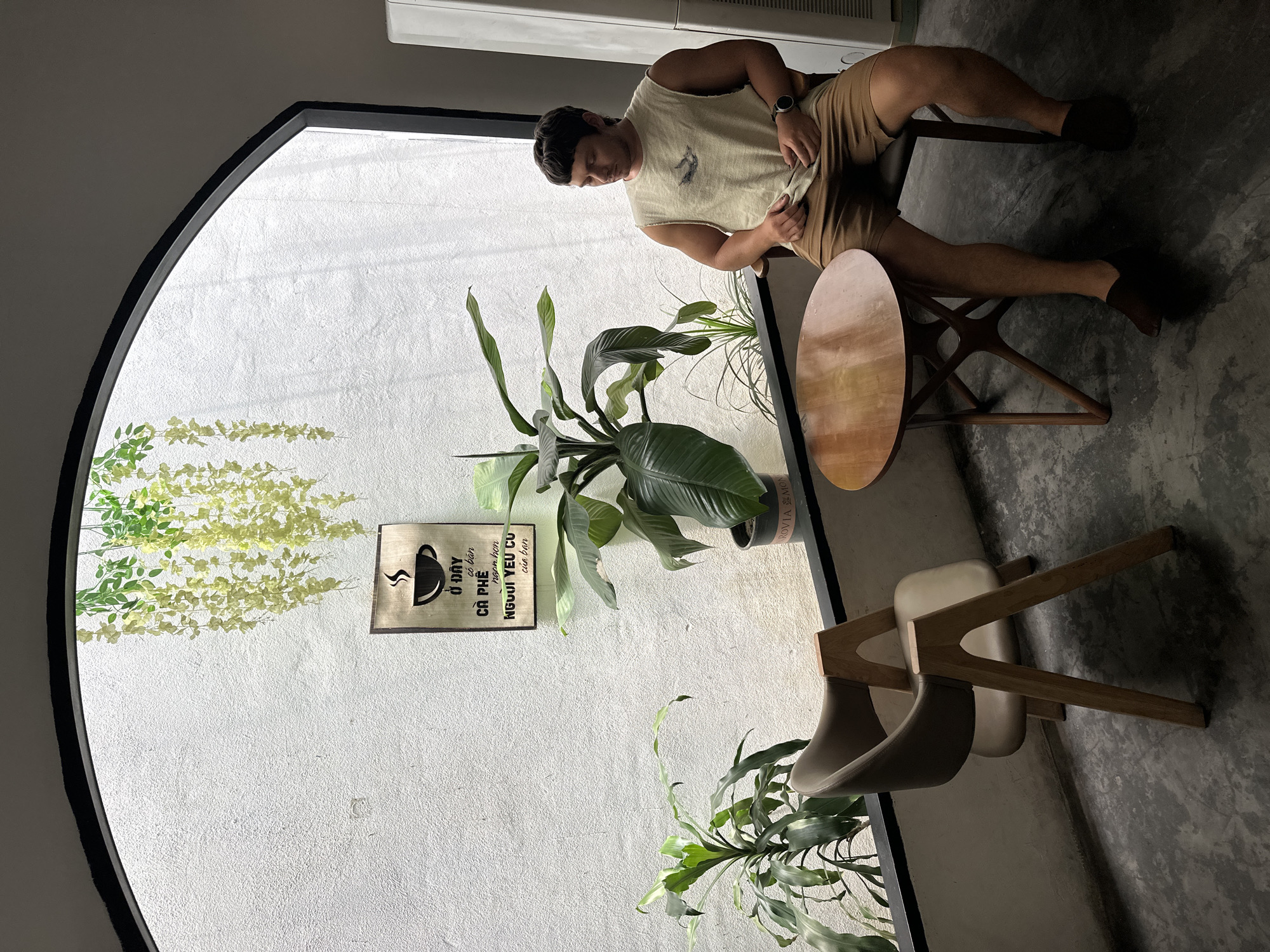 The image shows a man sitting at a small wooden table in what appears to be a cafe in Da Nang, Vietnam. He is wearing a light-colored sleeveless shirt with a graphic design and brown shorts. He is looking down at his hands, which are resting on his lap. Next to him is a grey chair with wooden legs. The cafe has a white textured wall with a large arched window, through which natural light streams in. Hanging plants and potted plants are visible, adding to the ambiance. A sign with Vietnamese text and a coffee cup graphic hangs on the wall. The floor is a dark, textured concrete. The overall scene suggests a relaxed and casual atmosphere.
