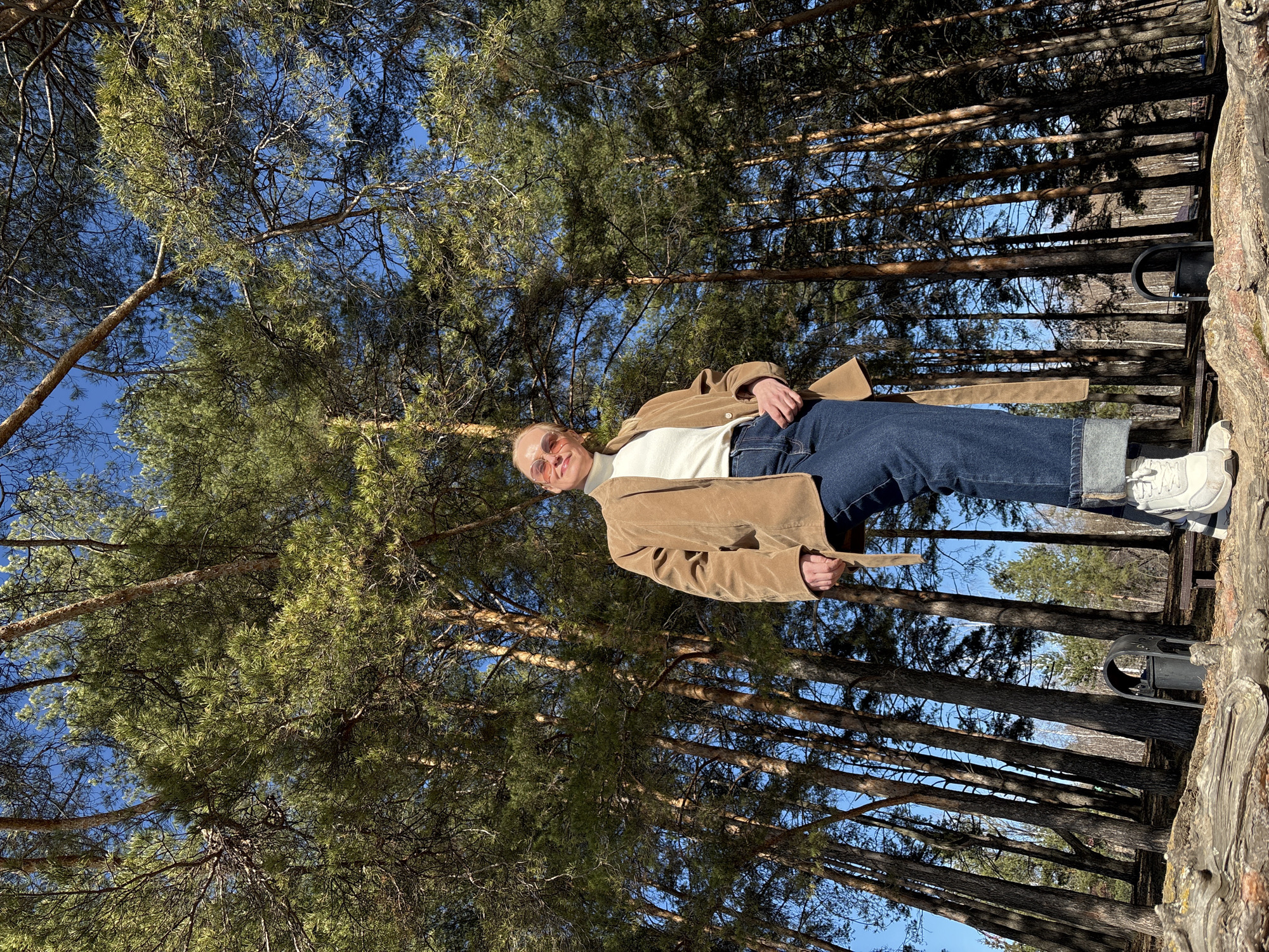 A young adult, possibly a woman, is captured in a full-body portrait set against a backdrop of towering evergreen trees and a clear blue sky. The subject is smiling broadly and looking directly at the camera, wearing round, orange-tinted sunglasses. They are dressed in a casual yet stylish outfit consisting of a light brown or tan corduroy coat, belted at the waist, over a white high-neck top. Dark blue, wide-leg or straight-leg jeans with cuffed hems and white sneakers complete the attire. The person's left hand is tucked into a pocket, while their right hand holds the coat's belt.
The setting is an outdoor forest, likely a pine or fir forest, characterized by numerous tall, slender tree trunks and dense green foliage that extends high into the frame. The sky is vivid blue and cloudless, indicating a bright, sunny day, possibly midday or early afternoon, given the distinct shadows cast by the trees. The person appears to be standing on a wooden structure, such as a platform or walkway, with a rustic wooden fence or barrier visible to their right. A dark, possibly metal, fixture is attached to this wooden structure. The scene evokes a sense of peace and contentment, with the subject enjoying the natural surroundings. No visible text is present in the image. The location context is Chaikovsky, Russia.