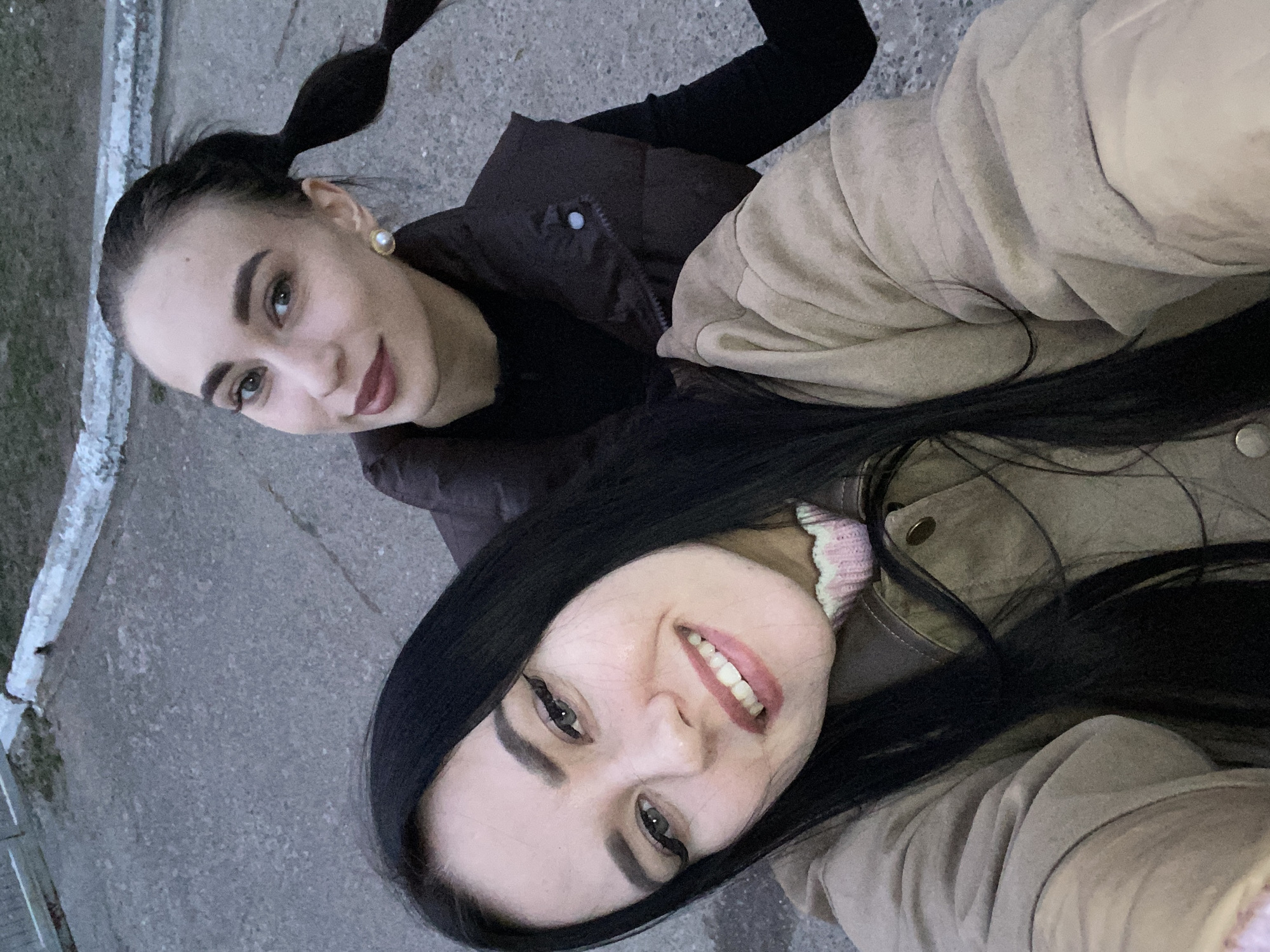 The image captures a close-up, horizontally oriented selfie featuring two young women in an outdoor setting in Chaikovsky, Russia.
In the foreground, a woman with long, dark, straight hair, parted down the middle, is smiling broadly with her teeth showing. She has prominent winged eyeliner and pinkish lipstick. She is wearing a light beige or light brown jacket, under which a collar with a light pink and white stripe or ruffle pattern is visible.
Slightly behind and above her, the second woman has dark hair styled in a distinctive bubble ponytail. She sports a subtle, closed-mouth smile, complemented by defined eyebrows and a darker reddish lipstick. She wears a dark brown or black quilted vest over a black long-sleeved top, and a large, round pearl earring is visible in her right ear.
The background consists of a grey, textured paved surface, likely asphalt or concrete, showing some faint cracks. On the left side of the frame, a low curb or wall with some white, possibly icy or painted, residue runs vertically. The lighting appears soft and diffused, suggesting either an overcast day or late afternoon/early evening, with no harsh shadows. Both women display pleasant and friendly expressions. No text is visible in the image.