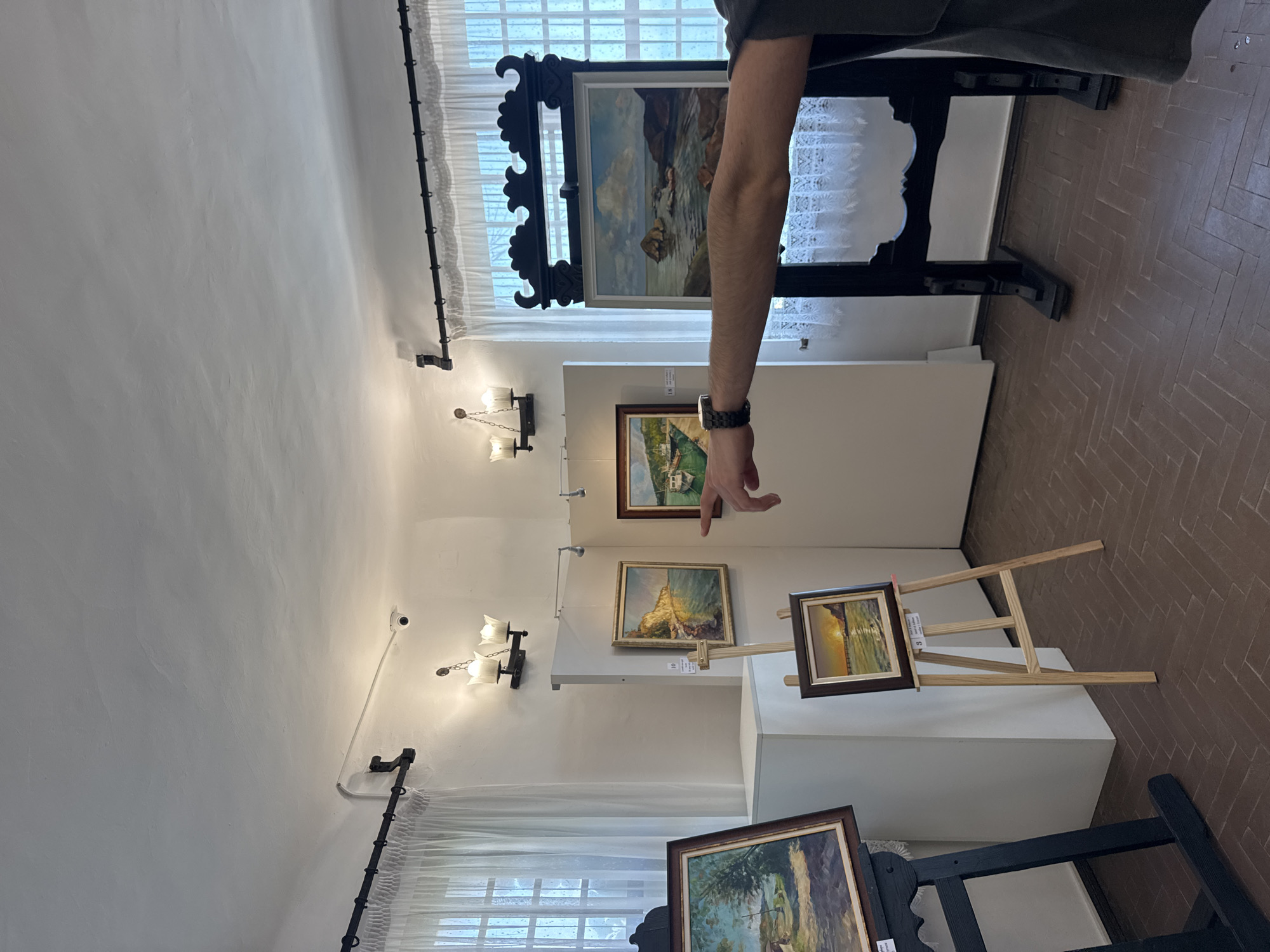 A person's arm points towards a collection of paintings displayed in a room with white walls and a parquet floor. The paintings depict various landscapes, including seascapes and coastal scenes. The room appears to be an art gallery or exhibition space, with art displayed on easels and on the wall. Two windows with white curtains are visible in the background. The lighting fixtures on the wall suggest it is an indoor space. The image is taken at the "Balchik Palace" in Balchik, Bulgaria, which was the summer residence of Queen Marie of Romania.
