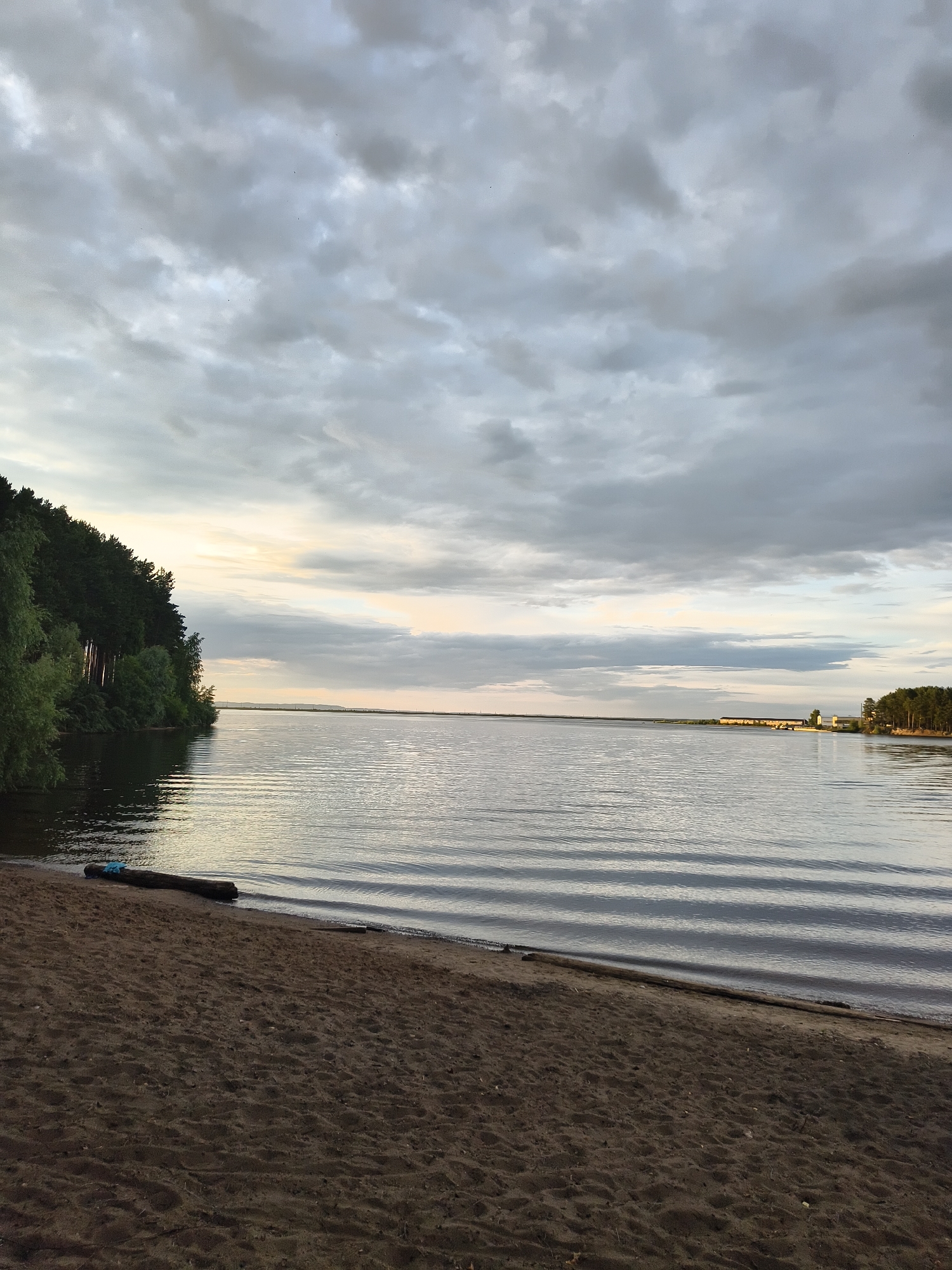 This media file captures a serene natural landscape, likely during the late afternoon or early evening, given the soft, diffused light. The scene is dominated by a vast expanse of calm water, likely a lake or wide river, reflecting the overcast sky and creating gentle ripples on its surface.
The left bank of the waterbody is lined with a dense forest of tall, dark green trees, creating a striking contrast with the lighter tones of the sky and water. A sandy beach occupies the foreground, with a fallen log lying near the water's edge, adorned with a blue piece of cloth or rope.
The sky is filled with heavy, gray clouds, suggesting an impending or recent rain shower, though the light filtering through them indicates it's not actively raining. In the distance, across the water, a narrow strip of land is visible, also covered in trees, and beyond that, hints of man-made structures, possibly buildings or a dam, can be discerned. The overall mood is one of tranquility and natural beauty, with a sense of solitude. No people are visible in the image, and there is no visible text. The location context places this scene in Chaikovsky, Russia.