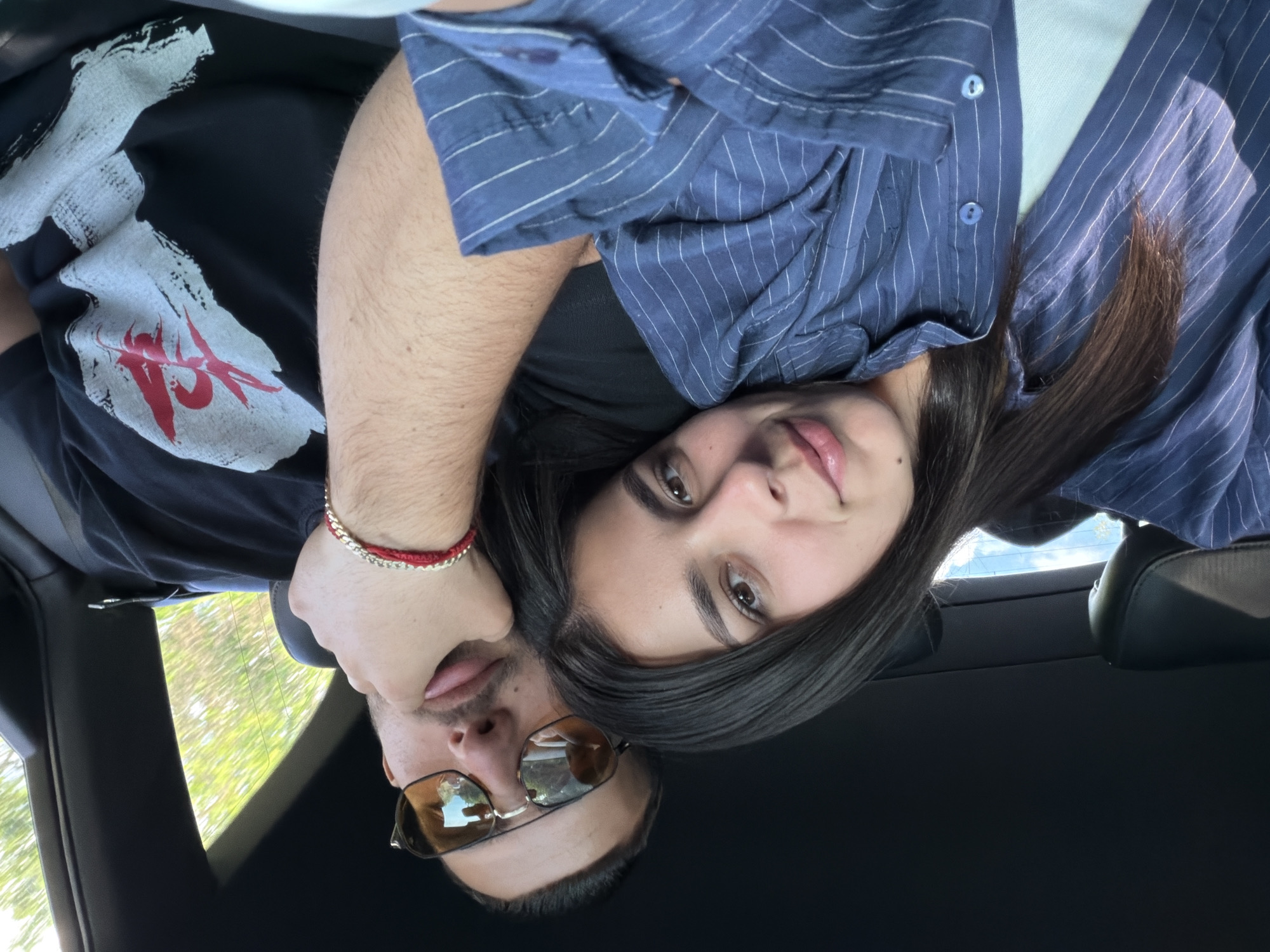 A selfie shows a couple in a car. The woman has long dark hair and wears a blue and white striped shirt. The man wears sunglasses and a black t-shirt with a white and red graphic. The car is black. Through the windshield, trees and the sky are visible. The location, Хаджийско, Bulgaria, is not discernible from the image.