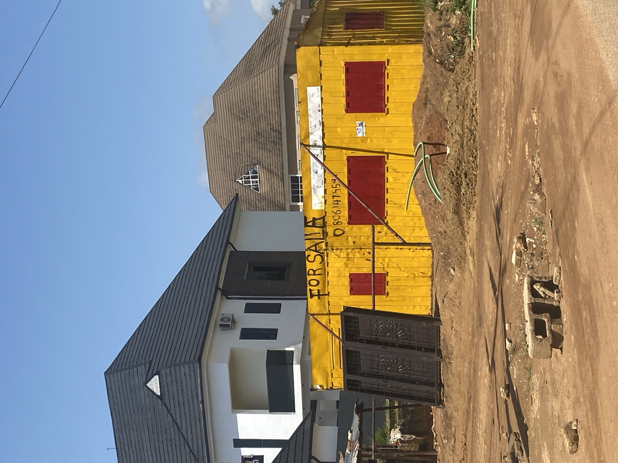 The image depicts a yellow shipping container converted into a structure, likely a shop or office, situated in Gura Top, Nigeria. The container is painted a bright yellow and has red-colored windows. Bold black lettering on the container reads "FOR SALE" and a phone number, "0806147559", is also visible. In front of the container, there are large metal doors, possibly part of an entrance, leaning against it. A pile of dirt and construction materials is present, with a green pipe snaking out from the pile. In the background, two modern houses are visible. One house is white with a dark grey tiled roof, featuring a balcony on the second floor. The other house has a brown tiled roof and a triangular window on its upper level. The sky is clear blue. The ground in the foreground is unpaved, consisting of dirt and scattered stones, suggesting an ongoing construction or undeveloped area.
