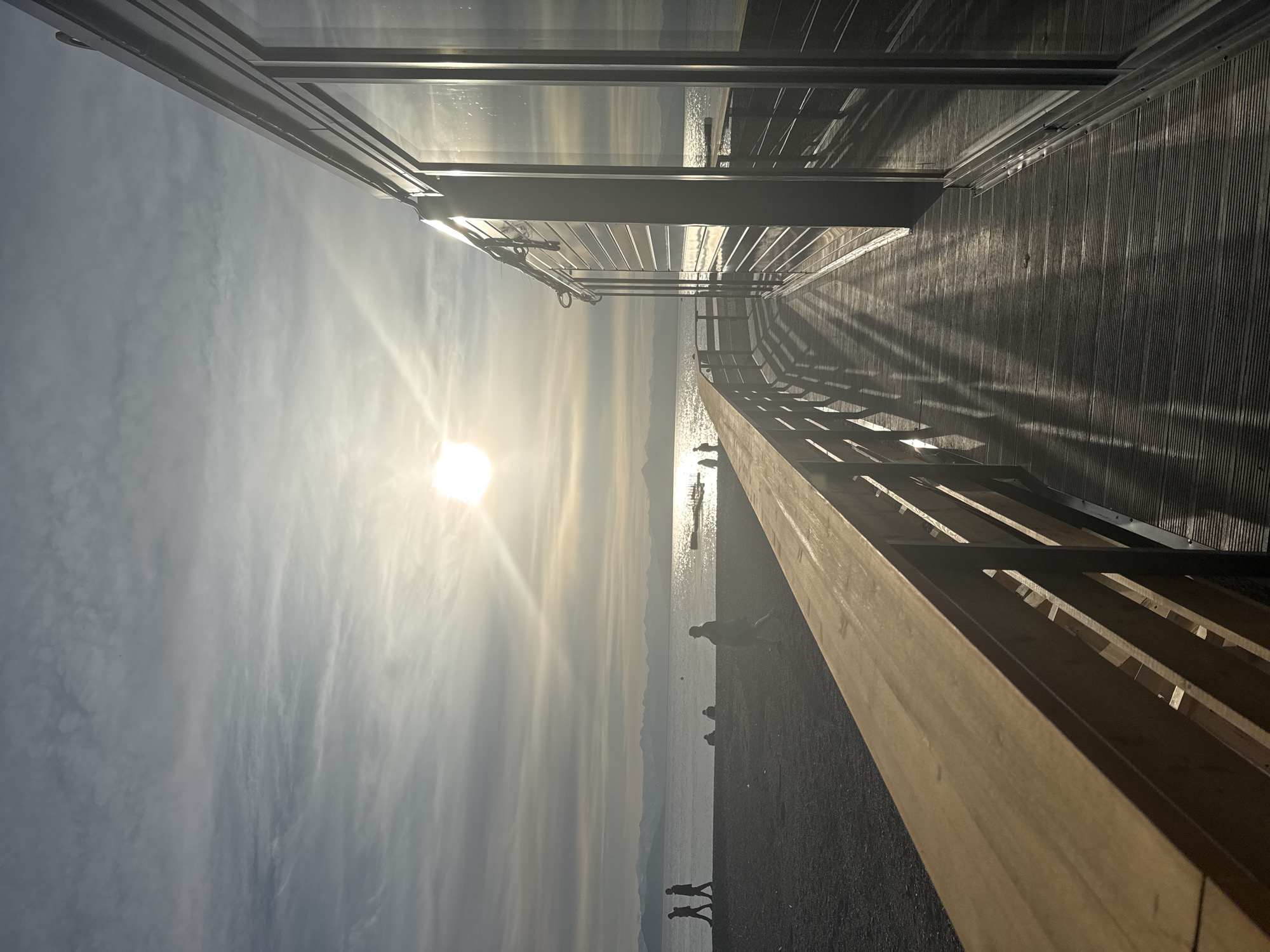This image depicts a waterfront scene, likely in Petropavlovsk-Kamchatsky, Russia, bathed in the warm, low light of the sun. The sun is positioned in the upper center of the frame, casting a bright glare and long shadows across the wooden boardwalk in the foreground. To the right, a modern building with large glass windows reflects the sky and surrounding environment.
The boardwalk extends along the water's edge, with a wooden railing bordering the left side, separating it from a dark, pebble-strewn beach. Several individuals are visible on the beach in the mid-ground, appearing as silhouettes against the water and distant mountains. Some are walking, while others are seated or standing.
The water itself stretches towards a hazy horizon, where snow-capped mountains are faintly discernible. A small boat or raft is visible on the water, catching the sunlight. The sky is partly cloudy, with wispy formations adding texture. The overall atmosphere suggests a serene, late afternoon or early morning setting.