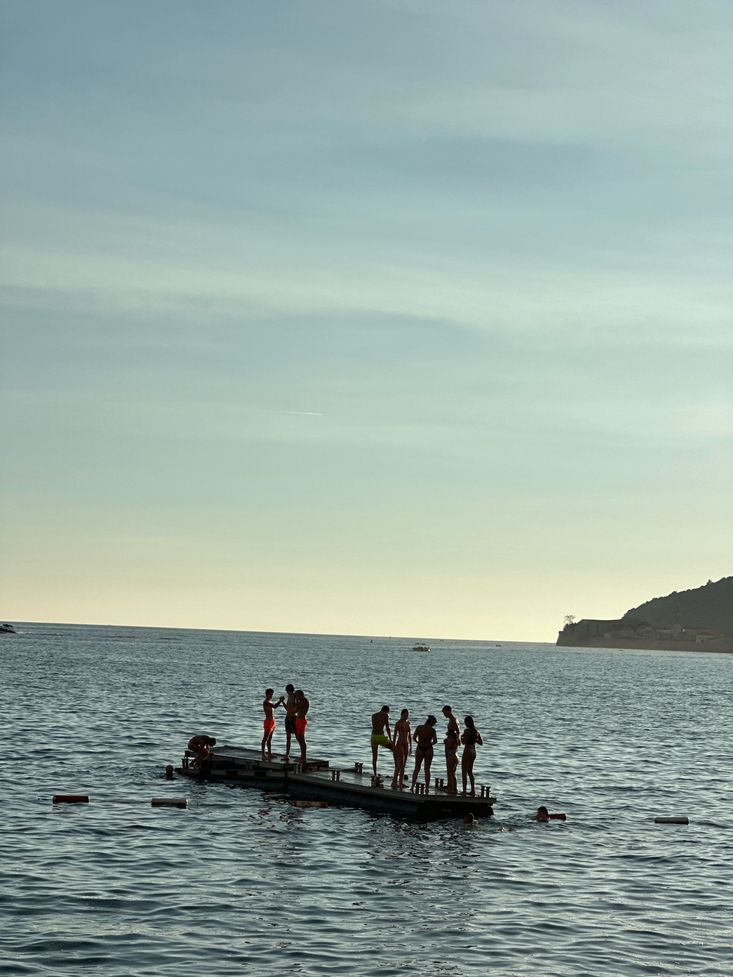 In Budva, Montenegro, a group of people are silhouetted on a floating platform in the sea. The sky is a pale blue with wispy clouds. The sea is a deep blue with gentle waves. In the background, a hilly coastline with some buildings and a tree is visible. Several boats are also present on the water. Some individuals are standing on the platform, appearing to interact, while others are in the water, swimming or wading. Red and white buoys are floating on the surface of the water, marking a swimming area. The scene suggests a recreational activity, likely during daylight hours with the sun low in the sky, casting long shadows.