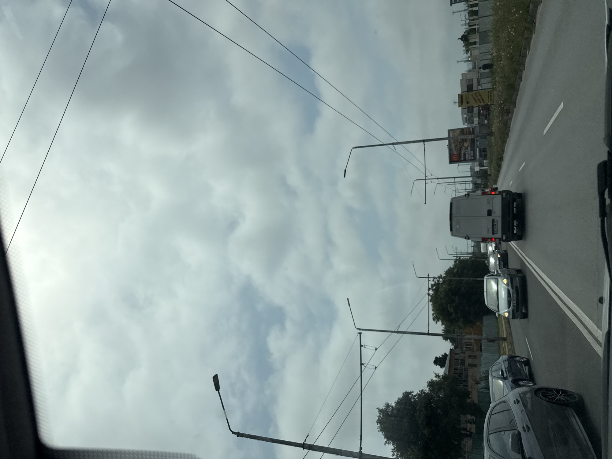 The image displays a roadside scene on an overcast day, captured from inside a vehicle. The perspective is unusually oriented, with a multi-lane road running vertically along the right side of the frame, occupied by several cars in what appears to be heavy or slow-moving traffic.
In the middle distance on the road, a white commercial van is visible, featuring a "BG" marking on its license plate, indicating Bulgaria. Behind it, a light-colored sedan and then a darker sedan are lined up. Part of another dark vehicle is in the immediate foreground on the bottom right. White lane markings are distinct on the asphalt.
To the right of the road, a barrier or fence separates it from a strip of sparse vegetation and various buildings in the background. Two notable billboards are present: a yellow one whose text is illegible, and directly below it, a clearer advertisement for "McDonald's" with the text "BIG TASTY".
The left side of the frame is dominated by an overcast sky, filled with grey clouds. Several utility poles or streetlights with an intricate network of overhead wires extend across the sky, characteristic of urban or suburban infrastructure. The scene depicts a daytime traffic situation in Varna, Bulgaria.