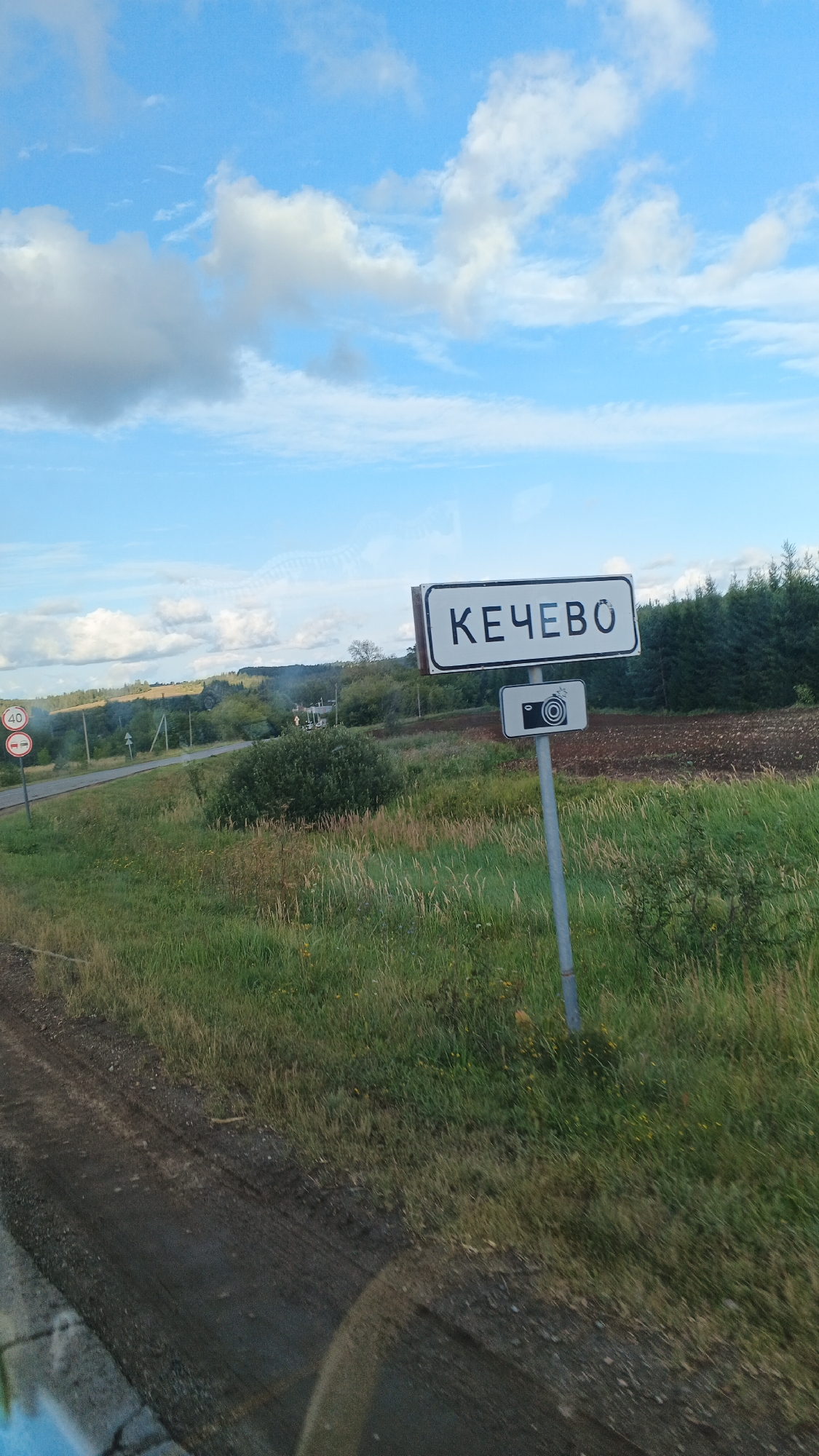 The image shows a road sign in the city of Кечёво, Russia. The sign, in white text on a black background, reads "КЕЧЕВО". Below it is a smaller sign depicting a camera symbol. The sign is mounted on a gray pole. The background shows a rural landscape with a road, fields, trees, and a blue sky with clouds. There are also other road signs visible, including a speed limit sign of 40. The photo appears to have been taken from a moving vehicle, as indicated by a blurred reflection in the lower left corner.