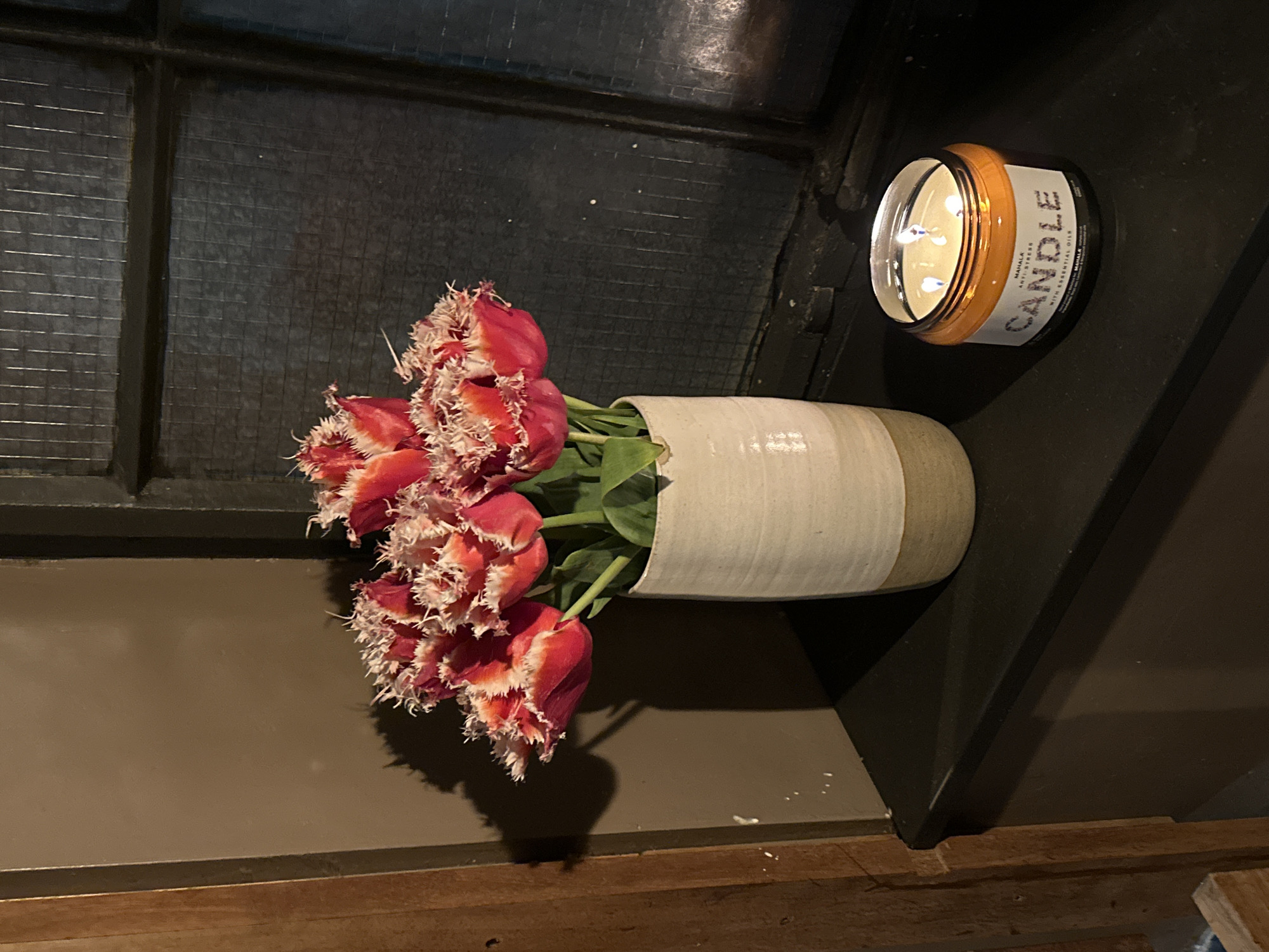 A dimly lit interior scene features a vase of red and white fringed tulips alongside a lit candle. The tulips are arranged in a cream-colored ceramic vase with a green base. The candle, labeled "MAHALK CANDLE," is in an orange container. The items are placed on a dark, possibly black, surface. Behind the flowers, a window with a grid-like pattern is visible. Based on the interior aesthetic, and the type of window frame it is impossible to say that this is in London, United Kingdom without further information.