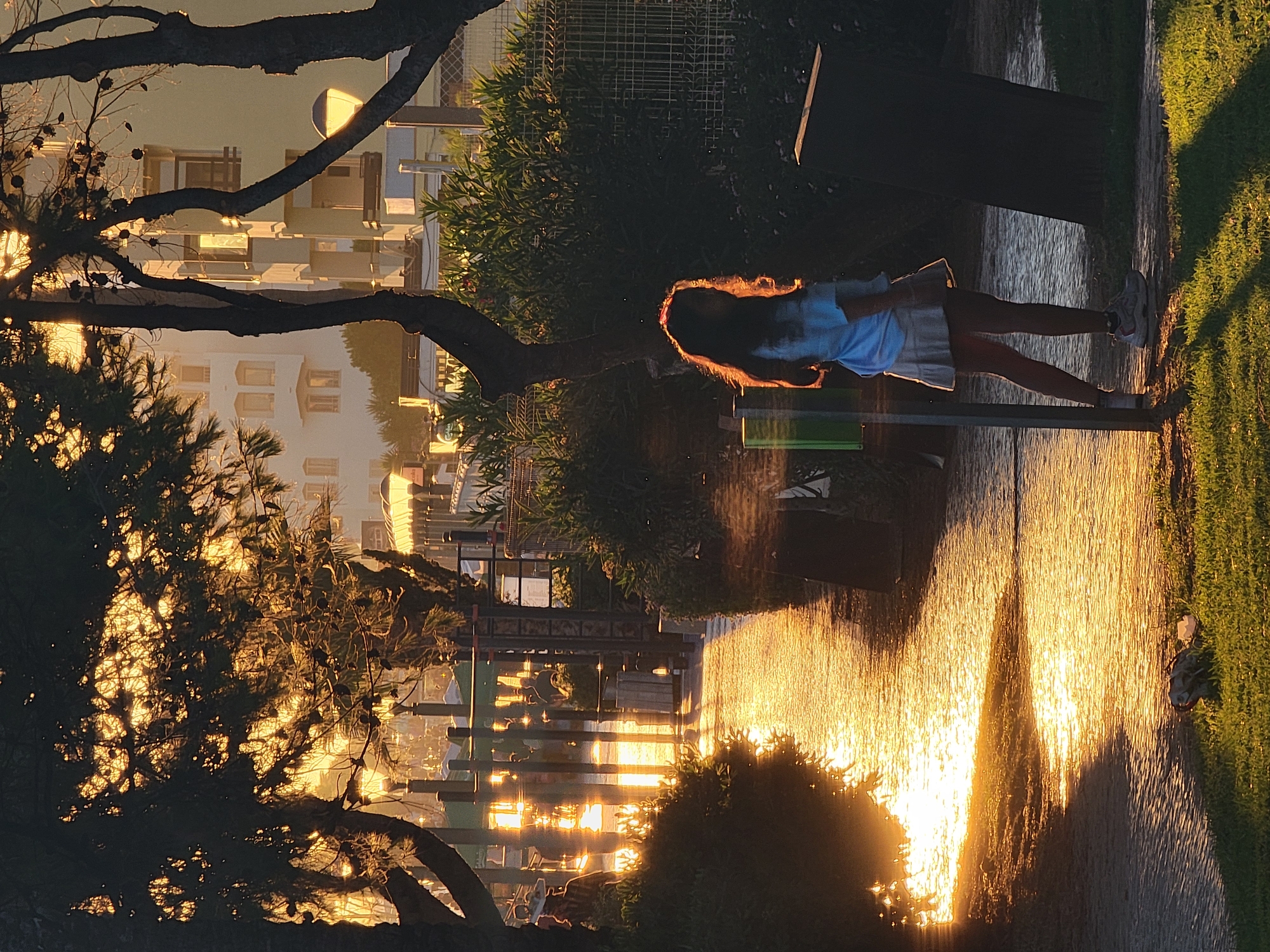 In Quarteira, Portugal, a young girl stands in the grass, bathed in the warm glow of the setting sun. She wears a light blue t-shirt and a white skirt, her hair illuminated by the golden light. The sun casts long shadows across a paved walkway where a golden reflection shimmers, suggesting the presence of water or a wet surface. Trees with dark branches and needles frame the scene, with pinecones visible. In the background, a building with multiple windows stands behind lush green foliage. Streetlights are visible along the walkway, and a fence marks the edge of the property. The overall atmosphere is serene, with the low angle of the sun creating a dramatic and beautiful play of light and shadow.