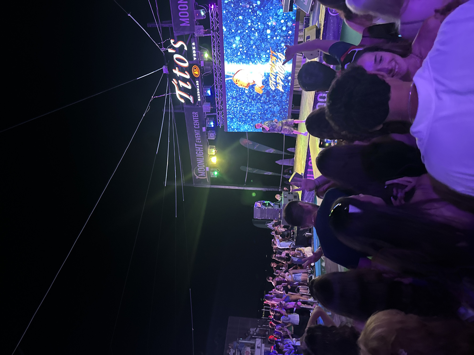 The image depicts an outdoor night event, likely a concert or party, at the Moonlight Event Center in Varna, Bulgaria. A stage is set up with a large LED screen displaying a graphic and text. On stage, a performer is speaking into a microphone. Above the stage, a neon sign reads "Tito's Handmade Vodka." A crowd of people is gathered in front of the stage, with some dancing and others observing the performance. The lighting is a mix of stage lights and ambient darkness. The overall atmosphere appears to be festive and energetic.