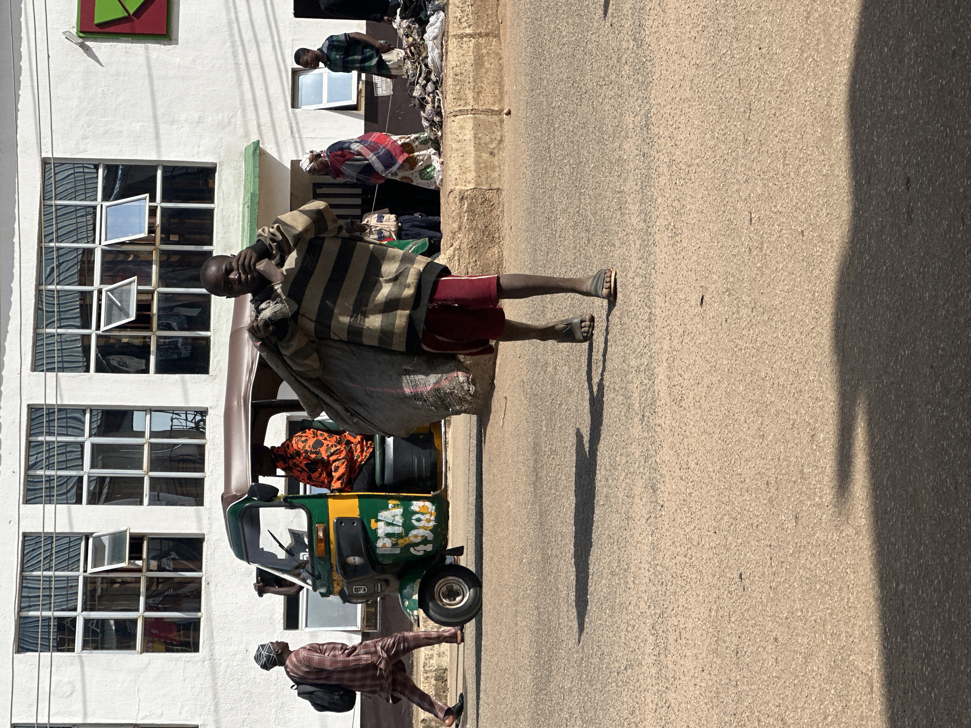 In Bukuru, Nigeria, a young Black male walks across a paved road carrying a large, dark grey, woven bag filled with what appears to be refuse or recyclables over his left shoulder. He is wearing a horizontally striped long-sleeved shirt with a hood, red shorts, and flip-flops. To his left, a green and yellow auto-rickshaw, also known as a "Keke Napep" or "Marwa", is parked at the side of the road. Inside the auto-rickshaw, a person wearing an orange and black patterned shirt is visible. Another Black male, wearing a patterned brown and white outfit and a dark cap, walks away from the auto-rickshaw towards the left, carrying a black backpack. In the background, a white building with numerous windows is present. To the right of the auto-rickshaw, a Black female wearing a colorful patterned wrap is standing, and further to the right, a Black male in a green and black striped shirt is visible. A pile of debris and what looks like discarded items is situated on the curb behind the people on the right. The scene is illuminated by bright sunlight, casting shadows on the road.