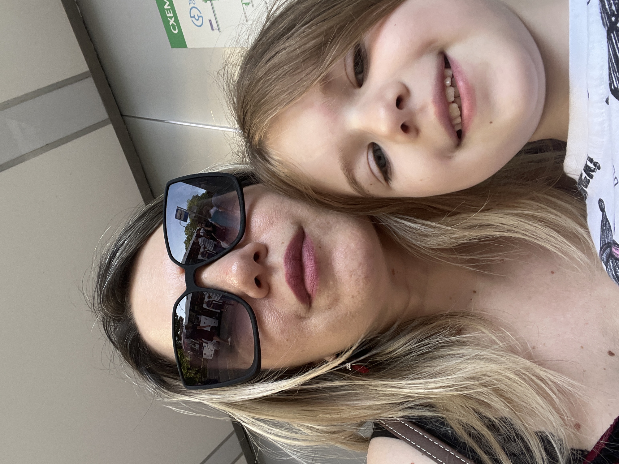 This media file is a close-up selfie of two people, an adult and a child, taken outdoors. The image is oriented vertically. The adult, on the left, wears sunglasses and has blonde hair. The child, on the right, has blonde hair and is smiling. In the reflection of the adult's sunglasses, a street scene is visible with people and trees. A sign with text and a diagram is partially visible in the background, on the upper left side of the image. The lighting suggests it is daytime. The general impression is a casual, happy moment shared between the two individuals. There are no explicit indications of weather, but the lighting suggests it is not overcast. There are no specific location cues within the image itself, but the prompt provides the context of Sochi, Russia.