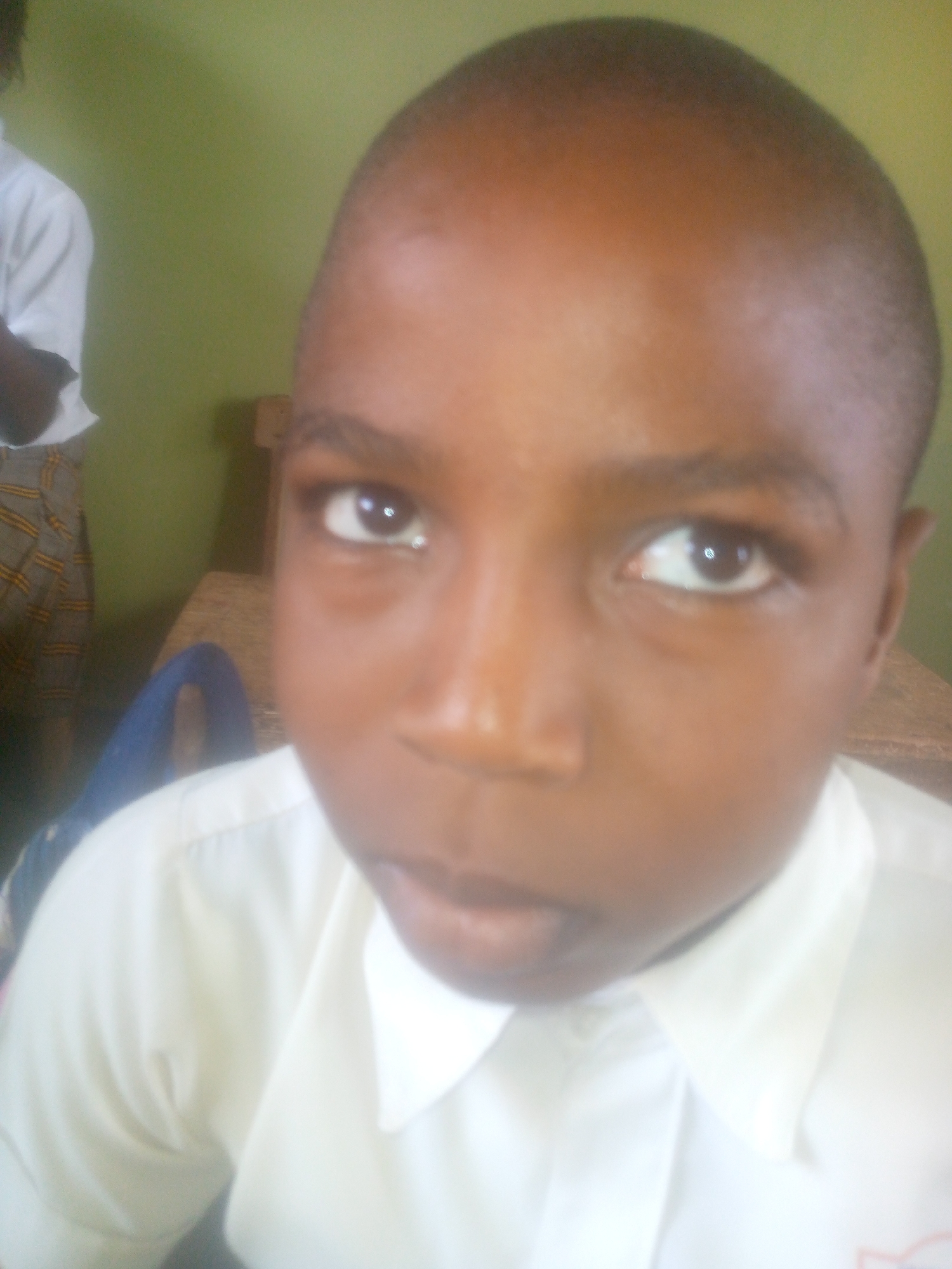 A close-up, slightly blurry, color photograph shows a young, dark-skinned boy with a shaved head wearing a white collared shirt. He is looking directly at the camera with a slightly open mouth. Behind him is a green wall. To the left, a partial view of someone in a white shirt and patterned skirt or shorts is visible. A wooden table is also partially visible in the background. The image appears to be taken indoors. The location in the city of Ikorodu, Nigeria is not discernable from the image itself.