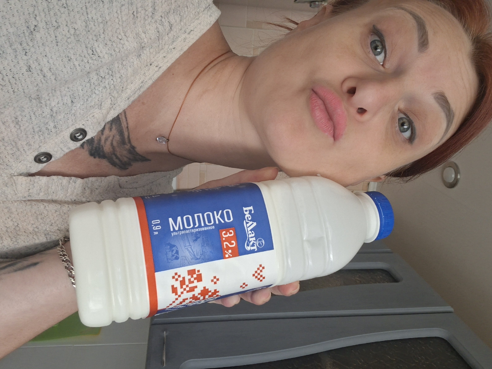 A woman with auburn hair and light green eyes is holding a bottle of milk. She is wearing a light-colored, long-sleeved shirt with buttons at the neck. She has on a delicate necklace with a clear pendant. A tattoo is visible on her upper chest and neck area. The bottle of milk has a blue cap and a label with Cyrillic text that reads "БЕЛАКТ" and "МОЛОКО", indicating it is milk. The percentage "3.2%" is also displayed prominently on the label. The background appears to be an indoor kitchen setting with gray cabinets. The overall scene suggests a casual moment captured at home. It is plausible this image was taken in Myadzyel, Belarus, given the Cyrillic text on the milk bottle and the prevalence of the "Belakt" brand in that region.