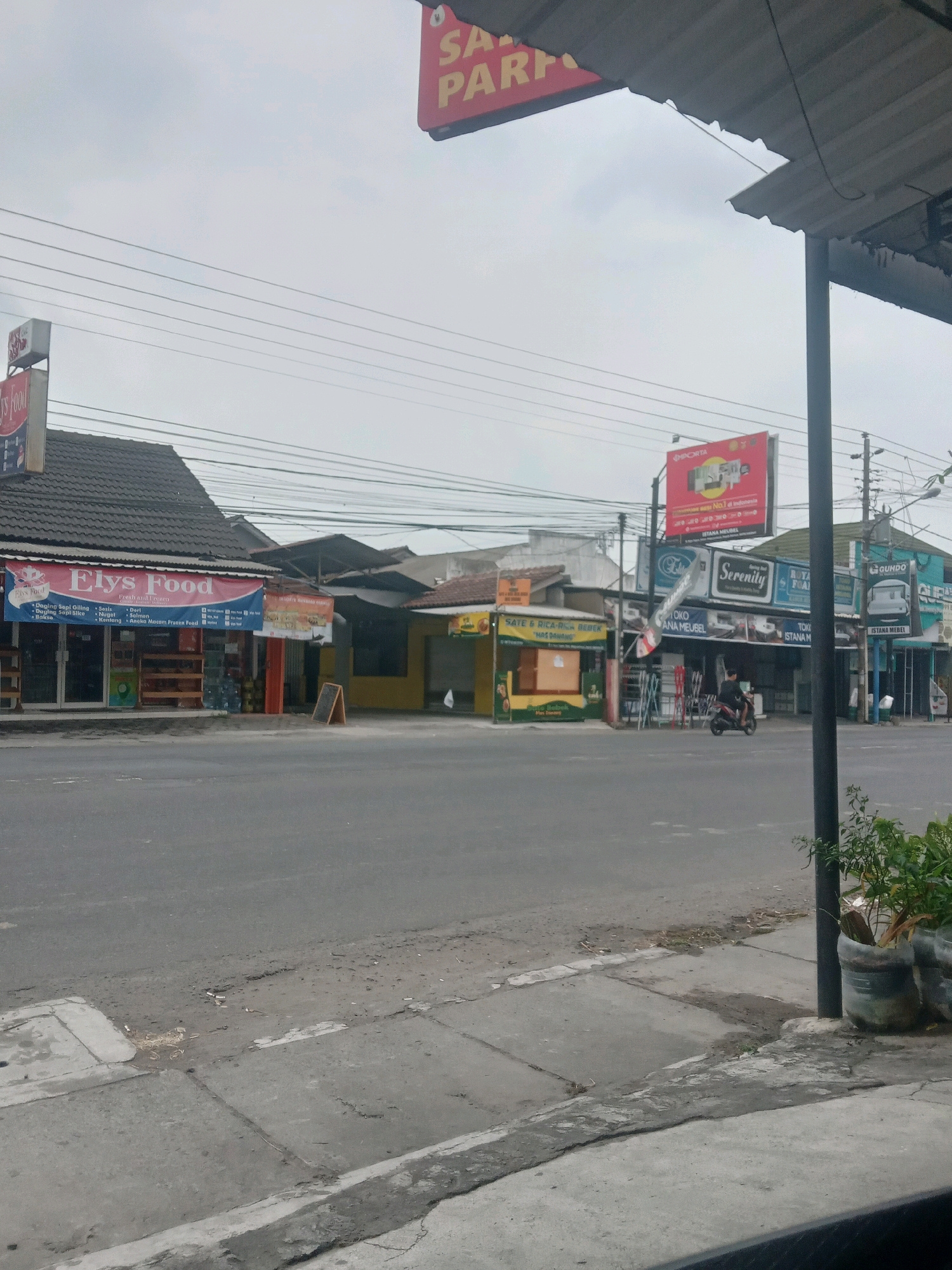 The image depicts a street scene in Maguwoharjo, Indonesia. The foreground shows a paved road and sidewalk with some debris and vegetation. In the middle ground, several shopfronts are visible, including "Elys Food," which appears to be a food store, and other businesses such as furniture stores. One shop has a banner advertising "SATE & BICA-RICE-BEBEK," suggesting it is a food stall. A person on a motorcycle is seen in front of one of the shops. Overhead, there are numerous electrical wires and a billboard. The sky is overcast. The overall environment suggests a commercial area in a town or city.