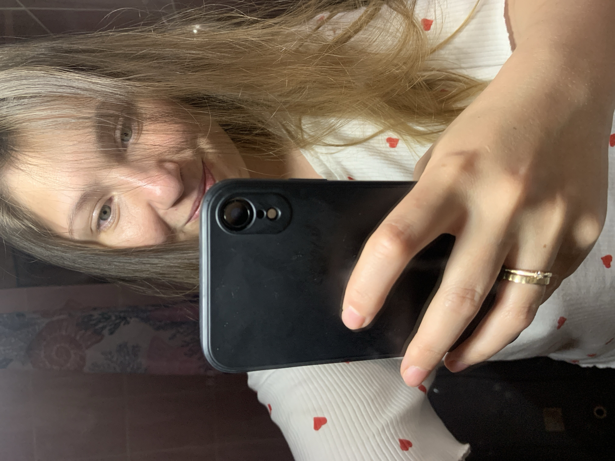 A person with light brown hair and light eyes is holding a black smartphone in their right hand, taking a selfie. The person is wearing a white, ribbed shirt with small red hearts printed on it. A gold ring is visible on the ring finger of the right hand. The background appears to be a room with brown and floral patterned elements. The location is specified as the city of Chaikovsky, Russia.