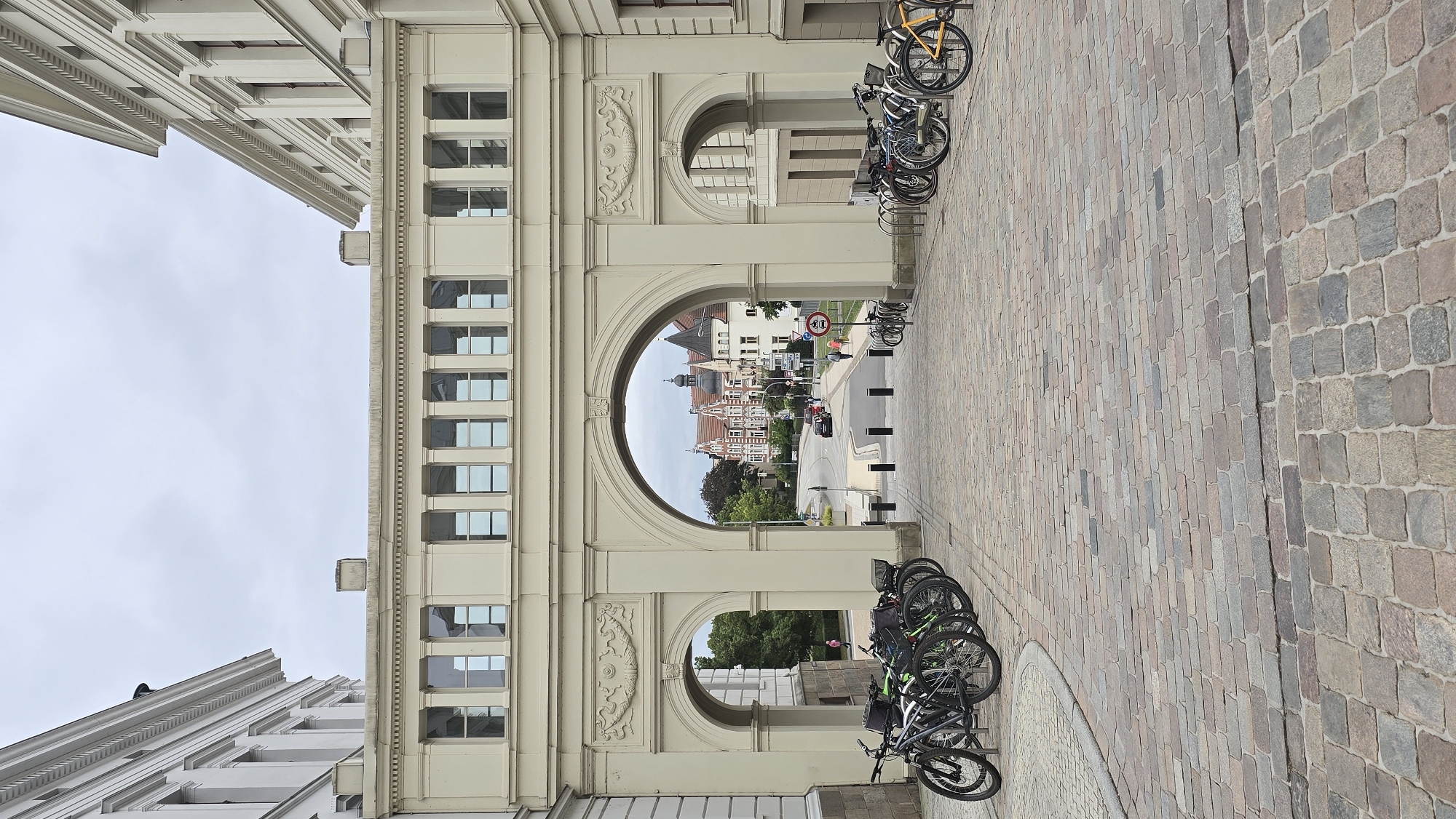 The image depicts an arched walkway connecting two classical-style buildings, likely part of a museum or administrative complex, in Schwerin, Germany. The walkway is supported by a large archway, allowing a view of a street with more classical buildings and trees in the distance. Several bicycles are parked on both sides of the walkway, indicating a mode of transportation commonly used in the area. The ground is paved with cobblestones, suggesting a historic district. The sky is overcast, contributing to a muted color palette. One person is visible walking on the street through the archway. The overall scene conveys a sense of architectural heritage and a peaceful urban environment.