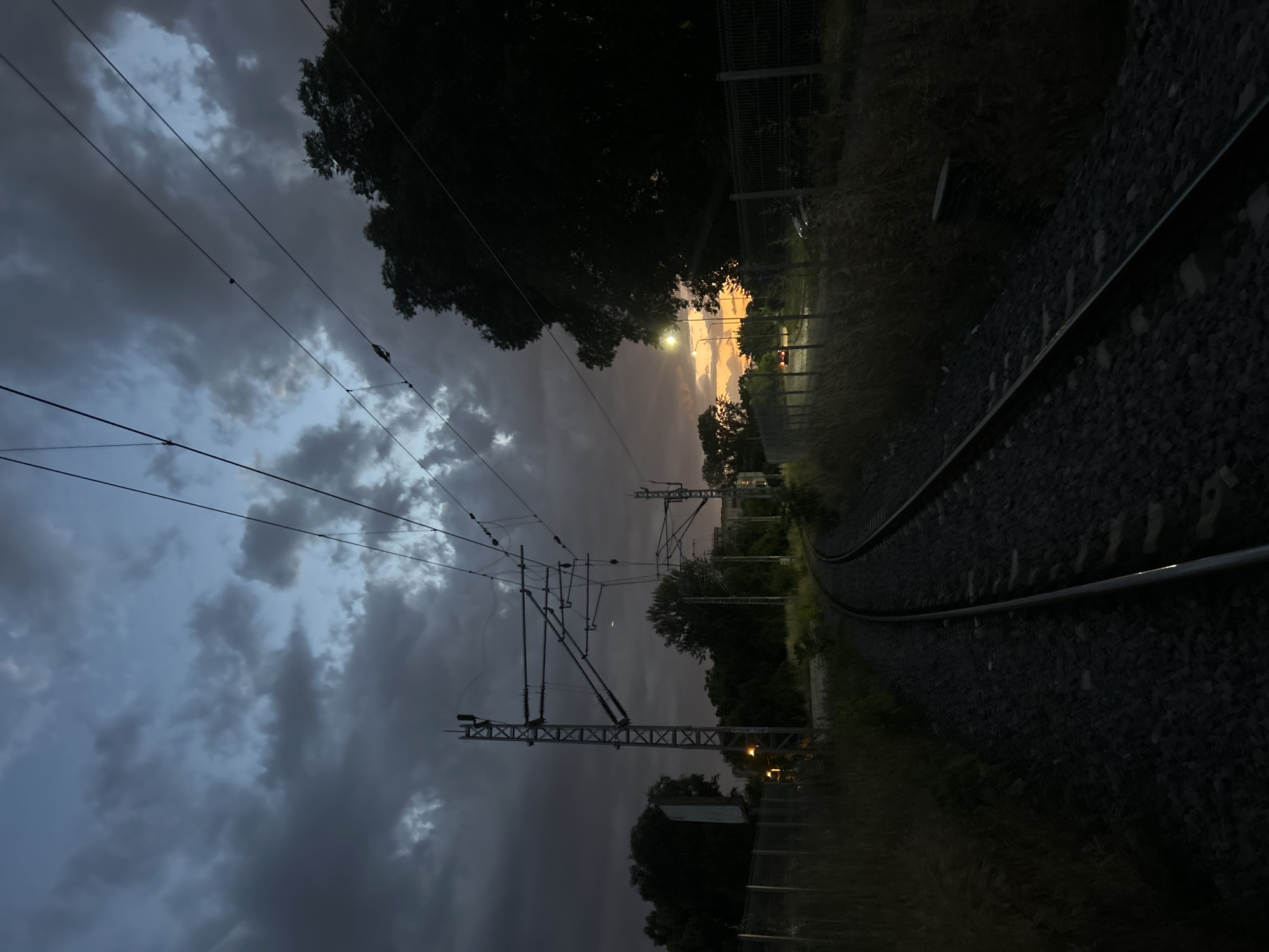 The image depicts a train track curving into the distance at dusk, likely on the outskirts of Plovdiv, Bulgaria. Overhead power lines are visible against a sky filled with dark, ominous clouds tinged with a faint orange glow from the setting sun. Trees line the edges of the tracks, and a fence runs parallel to the rails on one side. The rails themselves are dark and reflect a hint of light. The overall scene suggests a quiet, possibly abandoned, area. No people are visible.