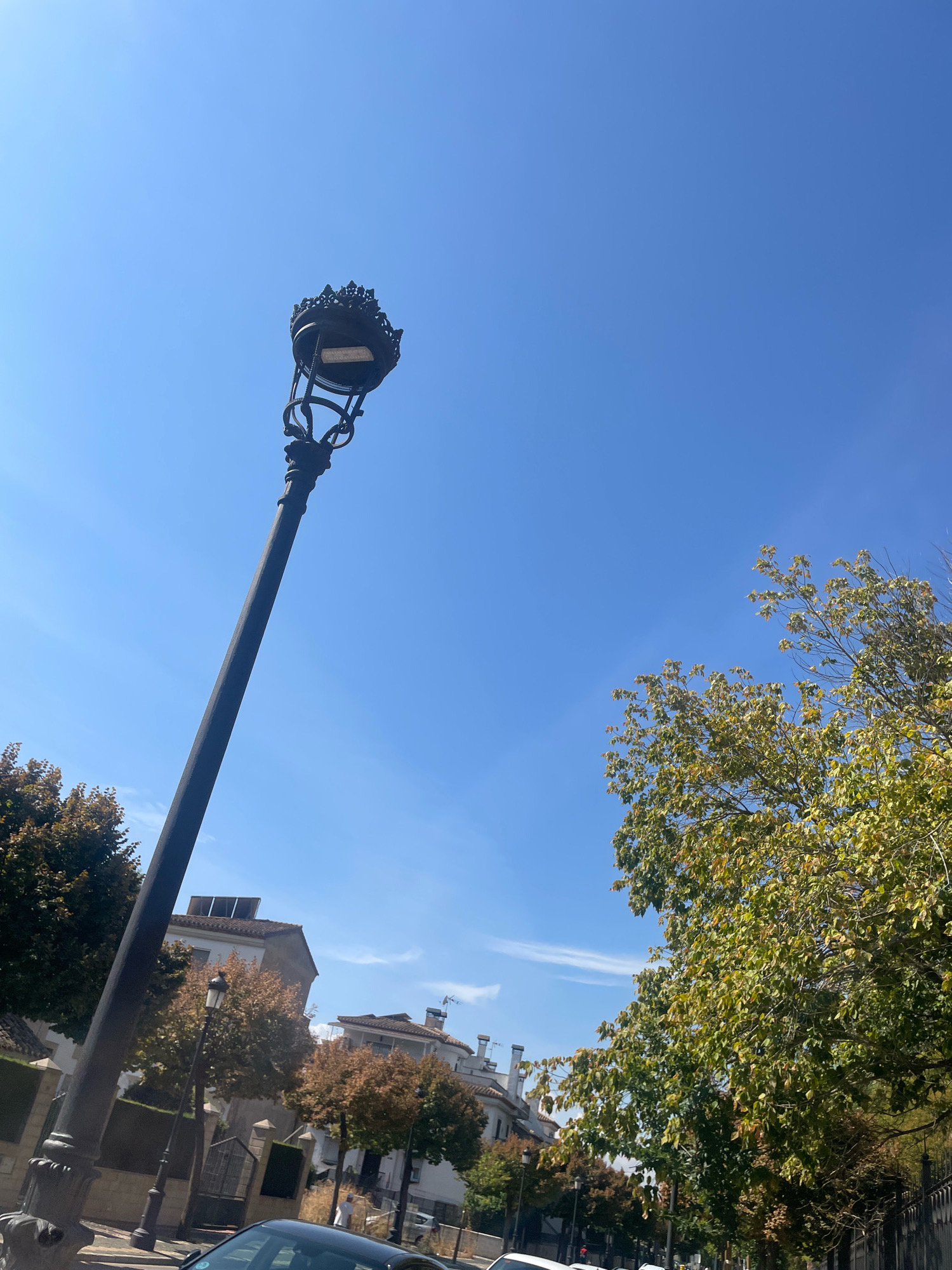 The image depicts a street scene in Atarfe, Spain, dominated by a tall, ornate black lamppost in the foreground, leaning slightly to the left. The lamppost features a decorative top resembling a crown. In the background, residential buildings with red-tiled roofs and white facades are visible, interspersed with trees displaying autumn foliage. A few vehicles are present on the street, and a person is walking in the distance. The sky is a clear, bright blue. The composition suggests a sunny day in a European urban or suburban setting.