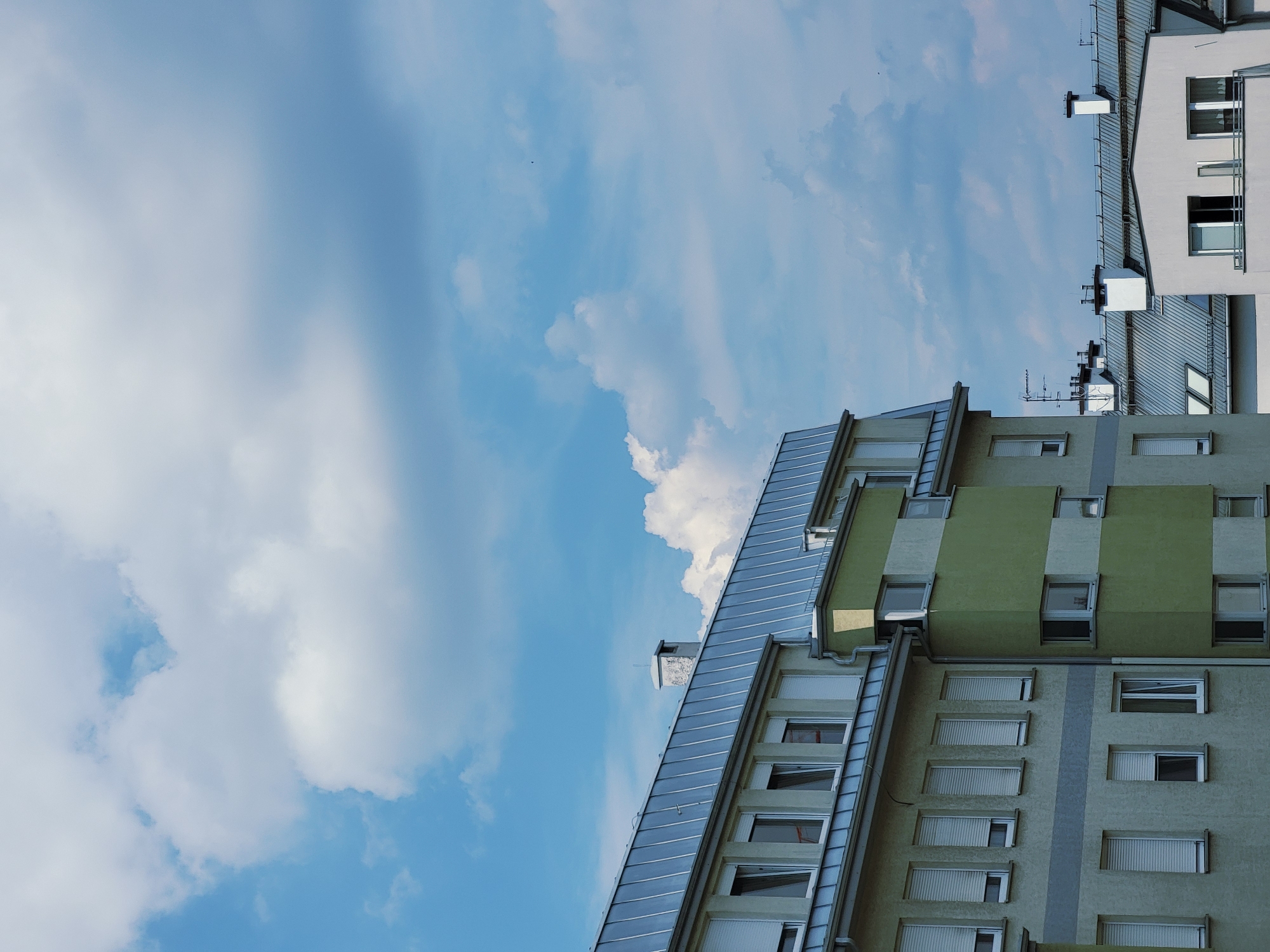 This is a photograph taken from a low angle, looking up at two adjacent apartment buildings. The building on the right is light gray with multiple stories and visible windows. It has a pitched roof and a series of chimneys. The building on the left is partially covered by a sloped, metallic roof that extends from its side. This building features a green facade with regularly spaced windows. The sky is a vibrant blue with scattered white clouds. The lighting suggests it is daytime. The image is oriented such that the buildings appear to be on the right side of the frame, with the sky dominating the left. The scene depicts an urban environment, likely a residential area. There is no text visible in the image. The photograph is taken from a perspective that emphasizes the scale and architecture of the buildings against the backdrop of the sky.