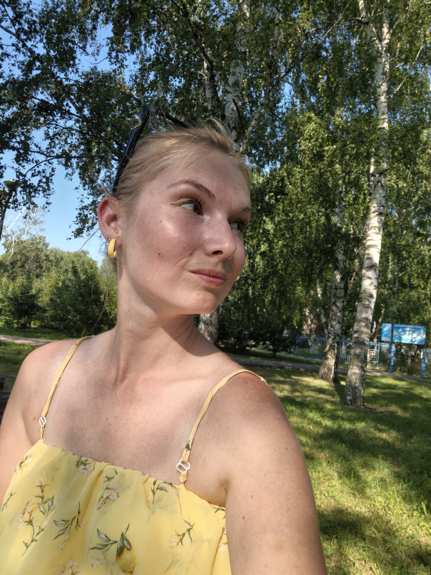 A young woman with blonde hair and a yellow floral dress is sitting on a park bench. She is looking to the right with a slight smile. Behind her are trees and a blue sky. The location is the city of Sterlitamak, Russia.