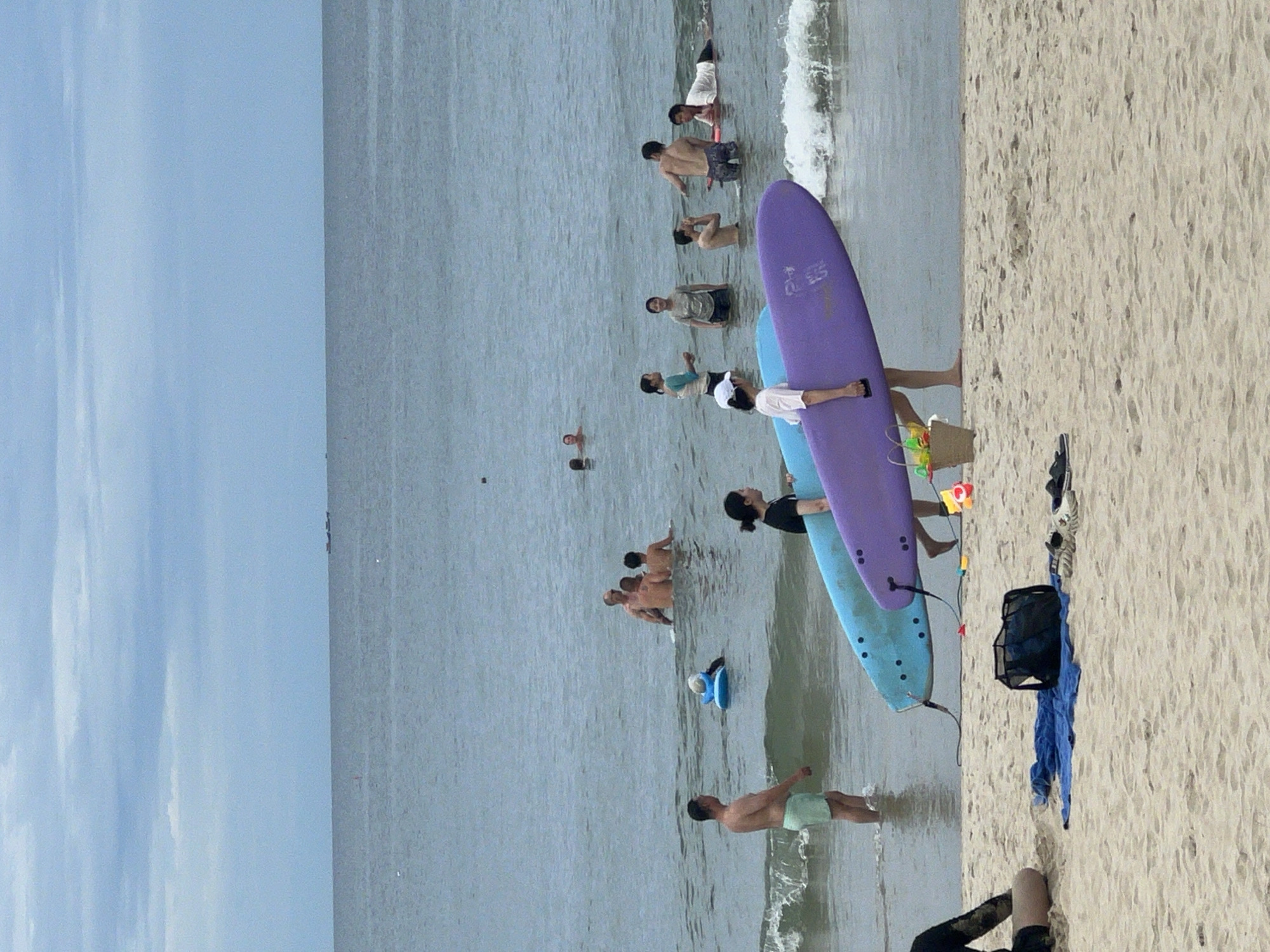 The image depicts a crowded beach scene in Đà Nẵng, Vietnam, likely during the daytime under overcast skies. The foreground is dominated by a sandy beach and the ocean, with several people engaged in various water activities. Two surfboards, one purple and one light blue, are prominently placed near the shore, suggesting surfing or a surfing lesson is taking place. Several individuals are in the water, some wading, some swimming, and others appearing to be learning to surf, with some lying on surfboards. The water appears calm with gentle waves lapping the shore.
On the beach itself, there are a few people, with some visible clothing and bags. The overall atmosphere seems relaxed and recreational, typical of a busy beach day. There are no clear indications of specific emotions, but the presence of multiple people enjoying the water points to a leisurely outing. The cloudy sky suggests it might not be a bright, sunny day, but it doesn't detract from the activity. The presence of surfboards and the general setting strongly suggest a coastal location, consistent with Đà Nẵng. No visible text is present in the image.