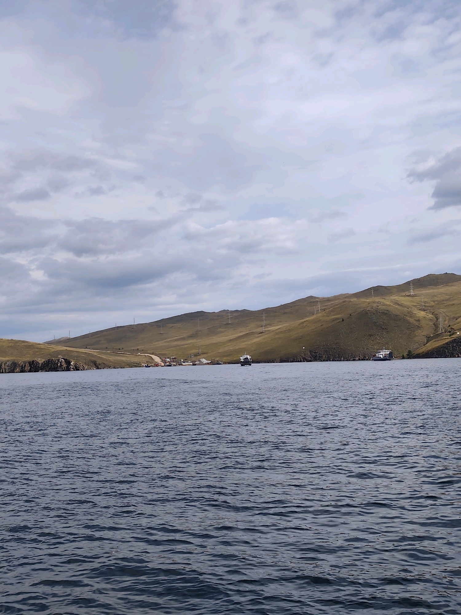 The image depicts a wide shot of a body of water, likely a lake or sea, with a distant shoreline. The water is a deep blue, with visible ripples and waves. The shoreline is characterized by rolling, golden-brown hills, with sparse vegetation. A dirt road winds its way up one of the hills. Along the base of the hills, near the water's edge, there appears to be a small settlement or village with several buildings and what looks like a small harbor. A few boats are visible on the water, including two larger vessels in the mid-ground and a smaller boat closer to the shore. Power lines and transmission towers are scattered across the hills, indicating some level of infrastructure. The sky is overcast with soft, grey clouds. The scene is devoid of any visible people or animals. The overall impression is one of a remote, natural landscape with a touch of human presence. The location is identified as Sakhiurta, Russia.