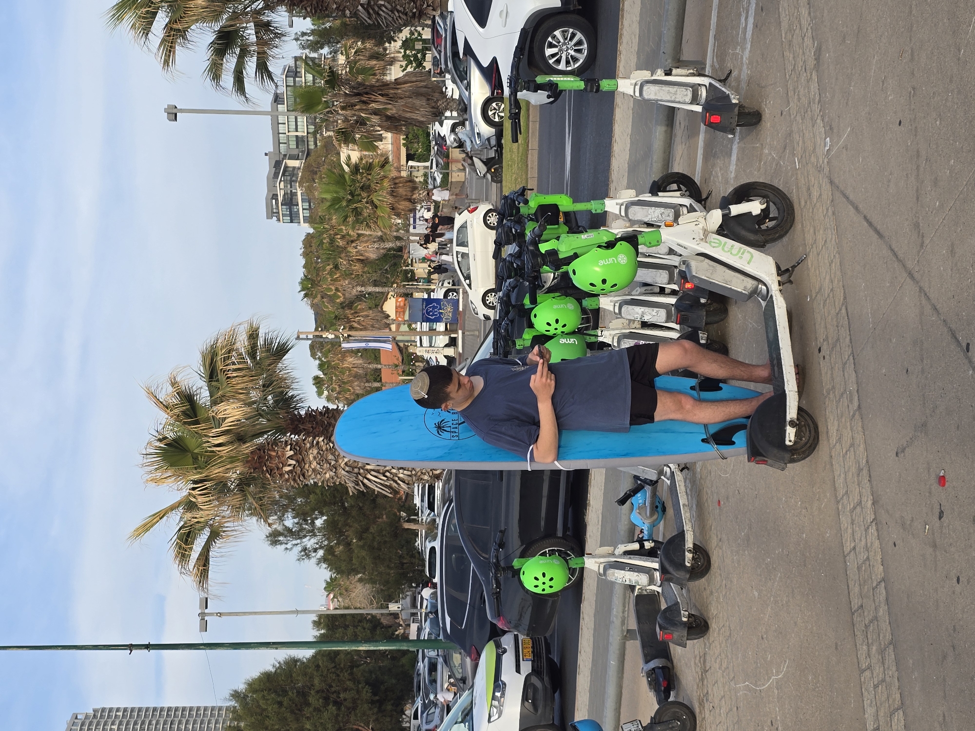 The image captures a street scene in Tel Aviv, Israel, characterized by a mix of urban and coastal elements. A young man wearing a yarmulke is lying on a light blue surfboard placed horizontally across a row of Lime electric scooters parked on the sidewalk. He is wearing a navy blue t-shirt and black shorts and is looking at a mobile phone he is holding. In the background, there are parked cars, including white, gray, and black models. Palm trees line the street, and buildings are visible in the distance, suggesting a location near the beach. The sky is pale blue, indicating daylight.