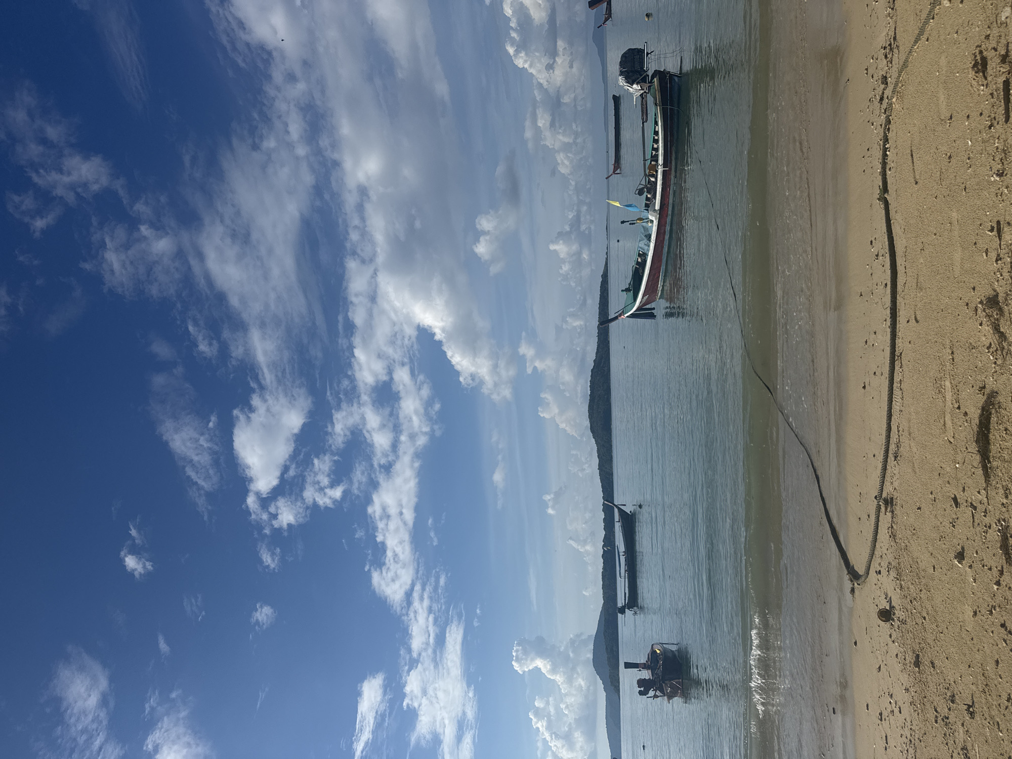 This is a photograph of the coastline at Rawai, Thailand. The image depicts a calm body of water with several longtail boats anchored offshore. The beach in the foreground is sandy and appears to be a calm day at sea. The sky is a clear blue with scattered white clouds. In the distance, lush green hills form the coastline. A thick rope lies on the sand, extending into the water, likely used for mooring the boats. The scene is serene and showcases the natural beauty of Rawai.
