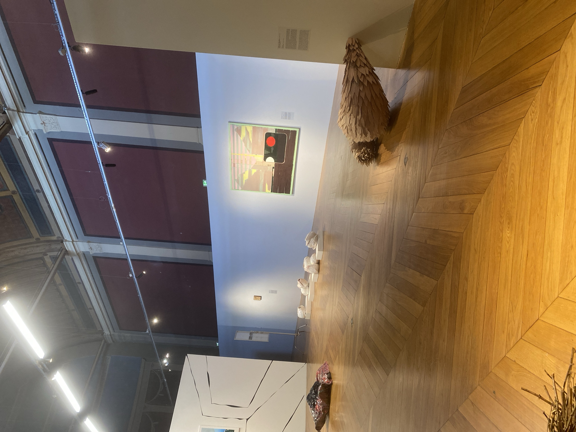 The image shows an interior view of an art gallery or museum, likely located in Paris, France. The room features light-colored wooden parquet flooring laid in a herringbone pattern. There are several art pieces on display, including a large, abstract painting depicting a traffic light, a tree-like wooden sculpture, a series of small white sculptures placed on white blocks, and a fractured white wall sculpture. The ceiling is high and architectural, featuring lights.