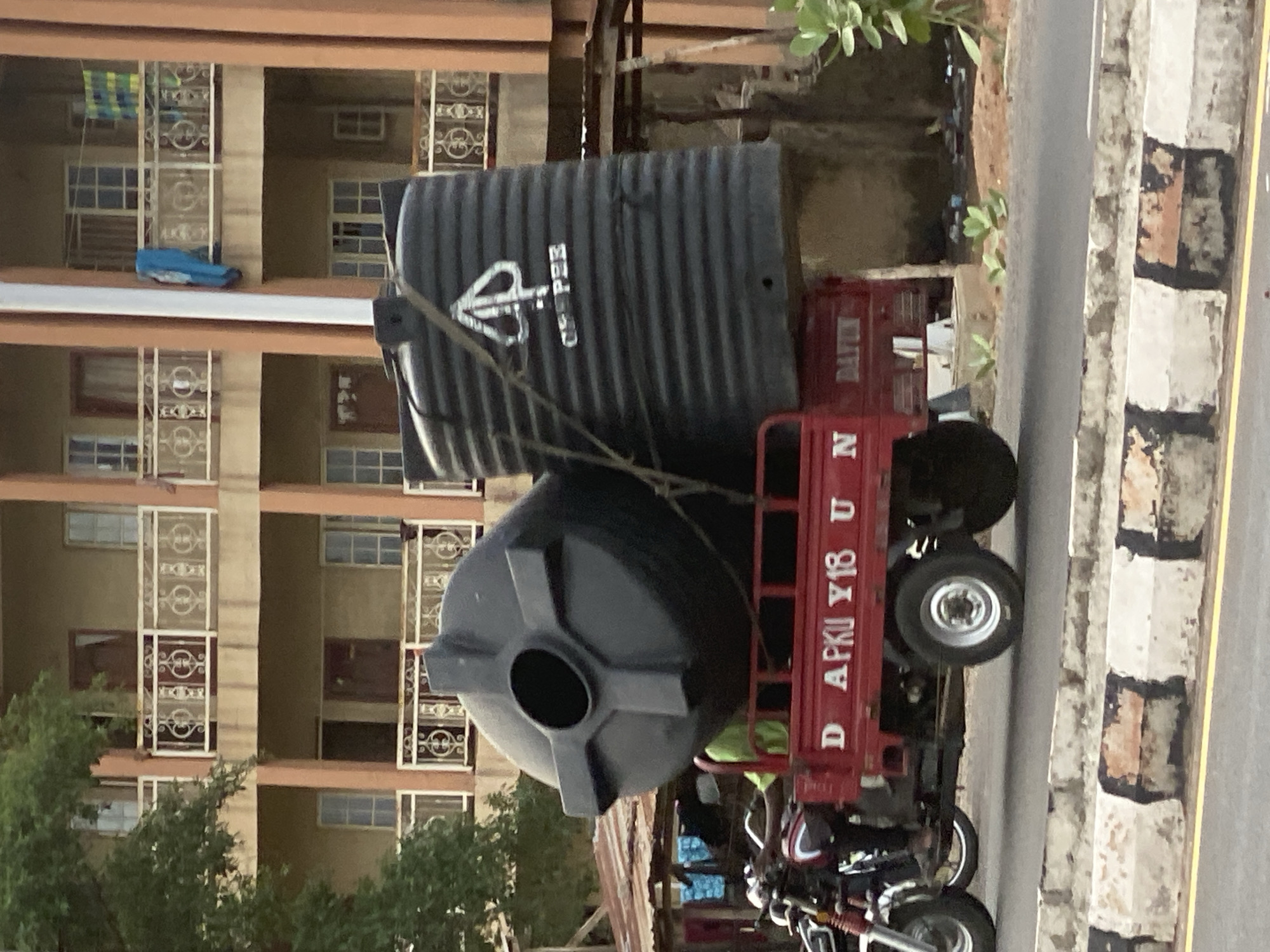 The image depicts a daytime scene in Zango-ndaji, Nigeria, centered around the transportation of water tanks. A red three-wheeled cargo vehicle, likely a motortricycle or a delivery tuk-tuk, occupies the midground. It carries two large, black plastic water storage tanks. One tank is cylindrical and lies horizontally, displaying the white text "OPEE" and a distinct logo. The other tank is more rounded and stands upright. Both tanks are secured to the vehicle with ropes. Visible text on the red vehicle includes "DATUN" and "N" on its side, and "DAPKU Y18 U" on its back. Portions of a motorcycle, presumably providing power to the cargo unit, are seen behind the tricycle.
In the background to the left, a multi-story residential building with light-colored walls stands prominently. It features numerous balconies, each protected by decorative white metal grilles. On one of the upper balconies, a blue object (possibly a folded cushion or mattress) and a colorful piece of fabric are visible. Green trees and foliage are situated in front of the building's lower levels. To the right of the vehicle, a paved road or street is visible, bordered by a curb painted with alternating white and black sections. A yellow line marks a lane on the asphalt. The scene captures an ordinary moment of goods transport and urban living.