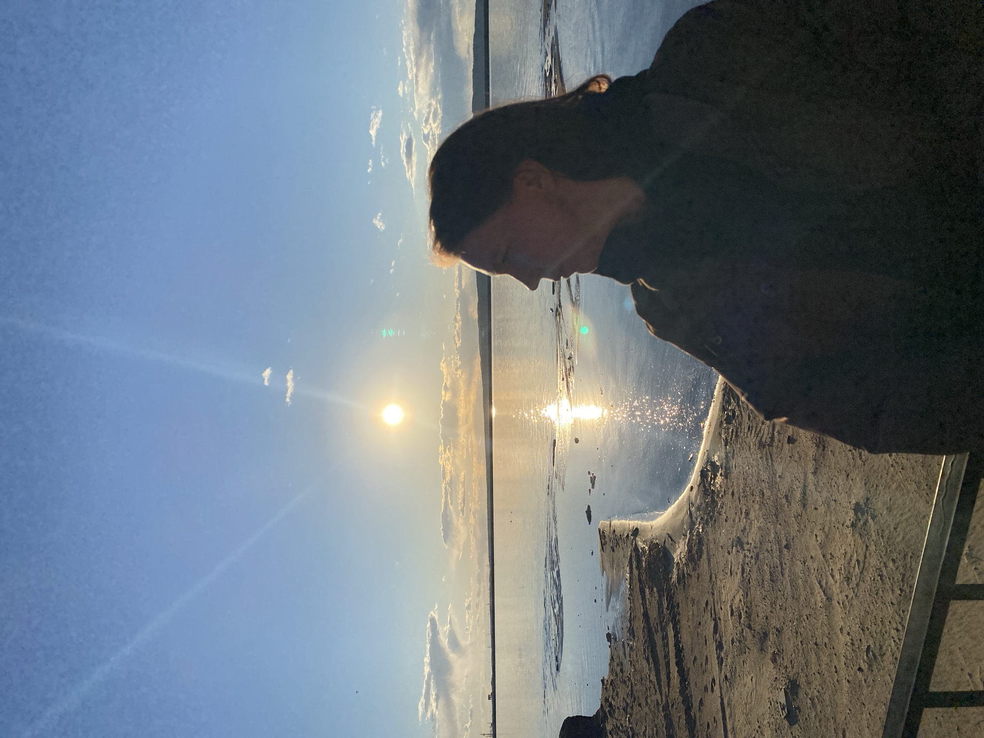 A landscape photograph, rotated counter-clockwise, shows a person standing near a body of water on a sunny day in what appears to be the city of Chaikovsky, Russia. The person, partially silhouetted, is positioned on the right side of the frame, facing downwards. To the left of the person, the sun is visible in the sky, and its reflection shimmers on the surface of the water. A shoreline is visible between the sky and the water, while a sandy surface is in the foreground.
