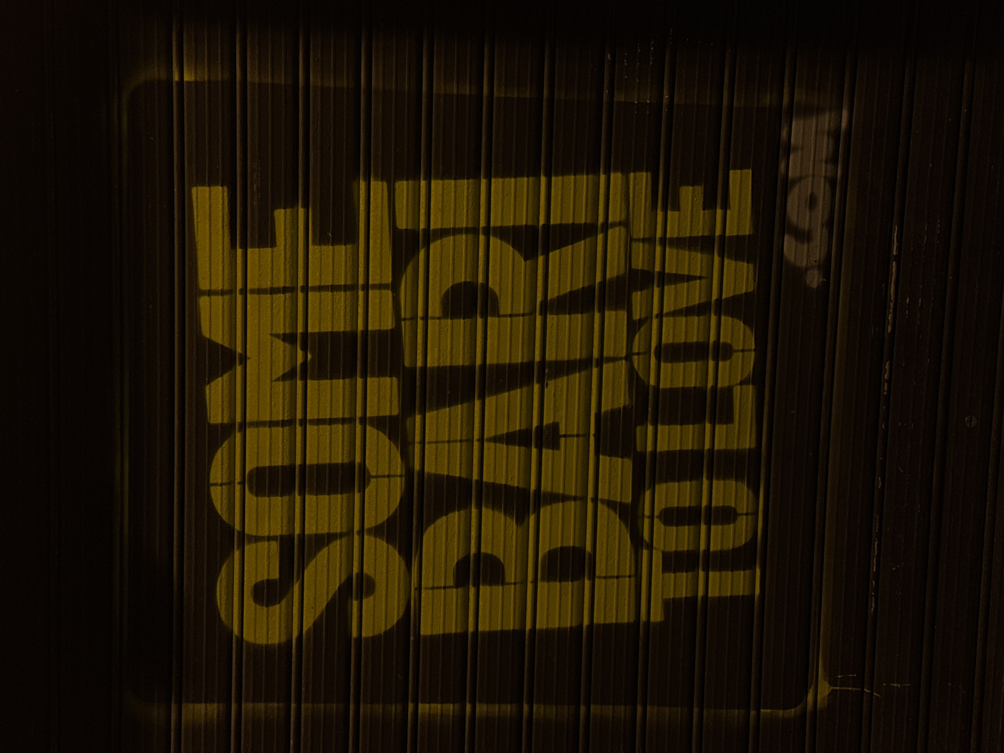 A nighttime photograph shows a sign stenciled onto a corrugated metal surface. The text, oriented vertically, reads "SOME BARI TO LOVE." The corrugated metal background is primarily dark, and the stenciled letters are a muted yellow-gold color. The letters are aligned to form a vertical rectangle. The word "BARI" appears in the center row, with "SOME" above and "TO LOVE" below. The background suggests an exterior location, perhaps a metal fence or a wall of a building. Based on the text, the sign is likely located in Bari, Italy.