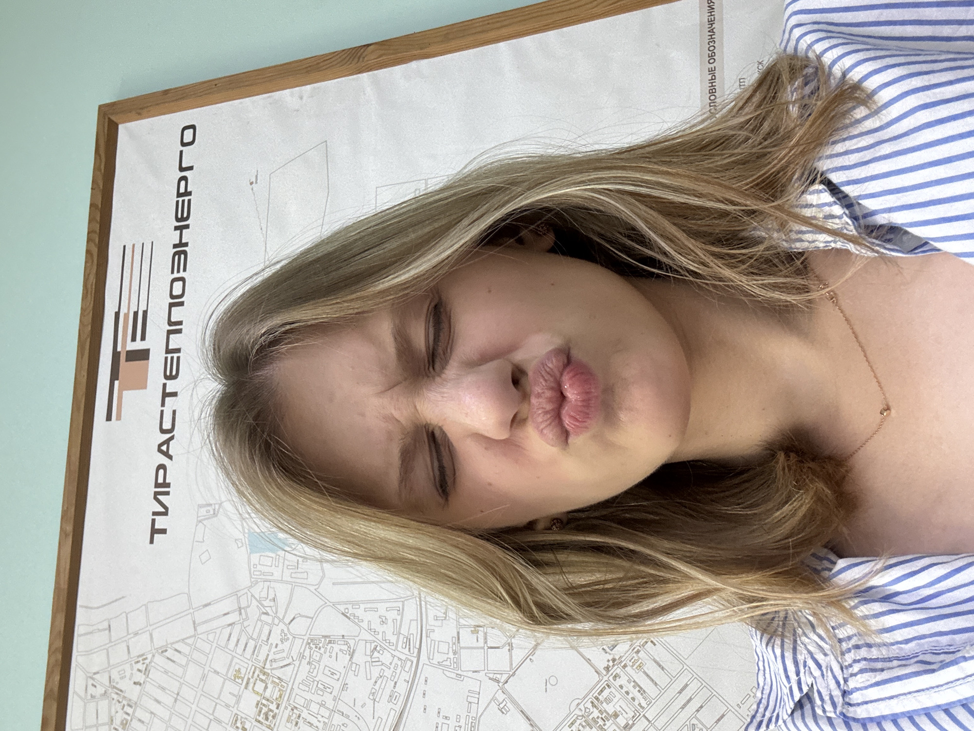 A young woman with blonde hair is taking a selfie in front of a framed map. She is making a puckered face with her eyes closed. She is wearing a blue and white striped shirt and a delicate gold necklace. The map behind her appears to be a city map, with street grids and building outlines. The text on the map is in Cyrillic script. A wooden frame surrounds the map, and the wall behind it is painted a light blue-green. It is impossible to determine the specific location in Tiraspol from the provided image.