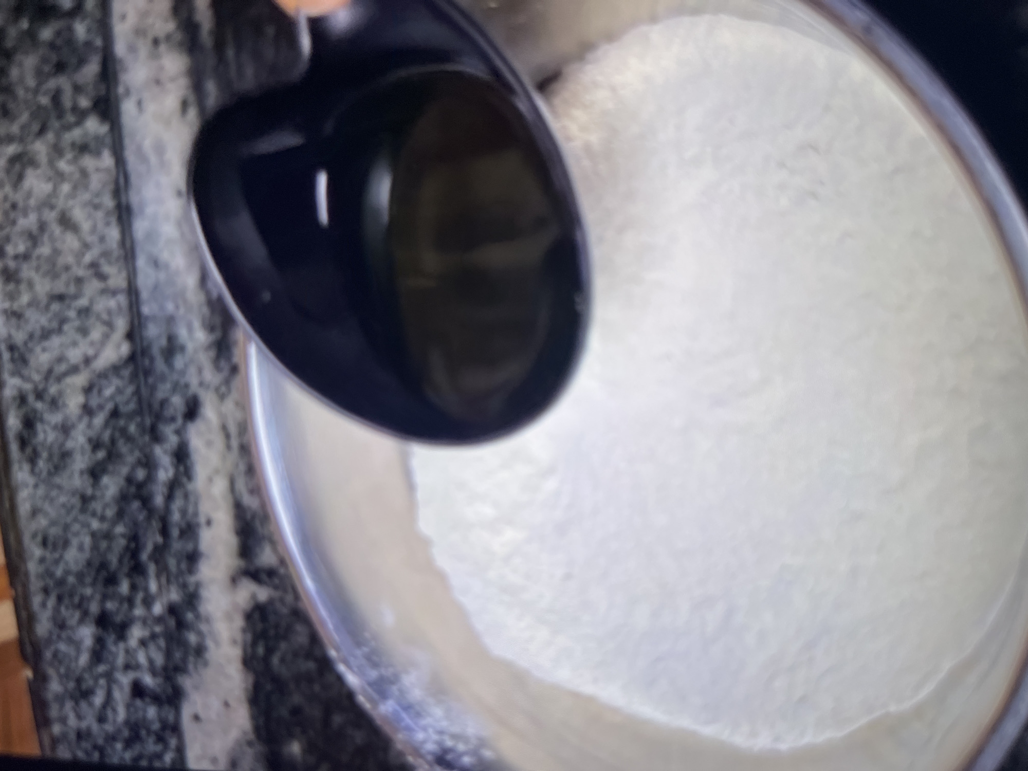 A dark ladle is held over a bowl filled with a white powdery substance. The ladle contains a dark, viscous liquid, likely oil. The bowl is metallic and sits on a countertop that appears to be made of granite or a similar stone material. The background is out of focus but suggests a kitchen environment. The image does not provide any information about the city of Abuja, Nigeria, or any specific location within it.