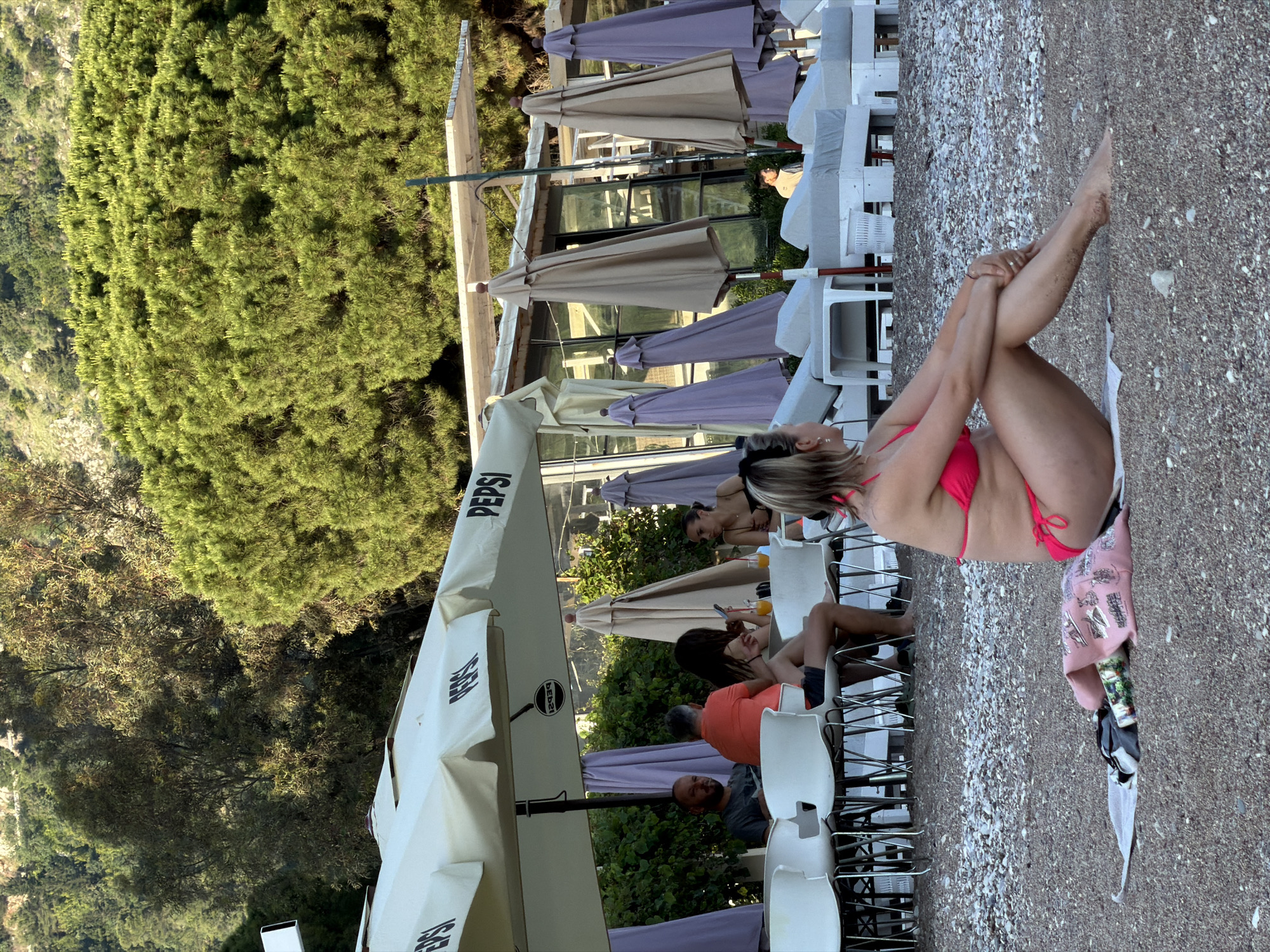 The scene is a sideways outdoor shot taken at a beachside bar in Budva, Montenegro. A fair-skinned woman in a pink bikini sits with her legs crossed and arms interlaced on a rocky beach. A white towel lies next to her, along with a discarded garment. Adjacent to the beach, a row of white plastic chairs and tables are arranged under numerous beach umbrellas, some with the “PEPSI” logo. Several patrons sit at the tables, including a woman in a black bikini and other individuals. Green trees can be seen in the background, suggesting the location is near a hillside or forested area. The overall lighting indicates it is likely a sunny day.