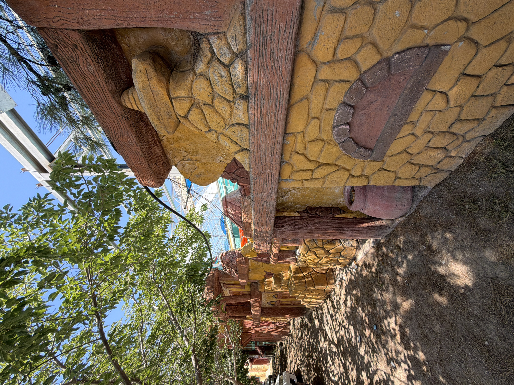 The image depicts an outdoor scene with a decorative wall made to resemble stone and wood. The wall is painted in warm tones of yellow and brown and features various textures and shapes, including what appears to be a stylized stone archway and a layered, conical structure. A large, reddish-brown clay pot sits on the ground next to the wall. Overhead, green tree branches with dappled sunlight cast shadows on the ground, which is covered in dirt and dried leaves. In the background, a white car is partially visible on the left, and a glimpse of a structure with a metal roof and blue sky can be seen. This scene is located in Kemer, Turkey, likely a tourist area given the decorative style. There are no people visible in the image.