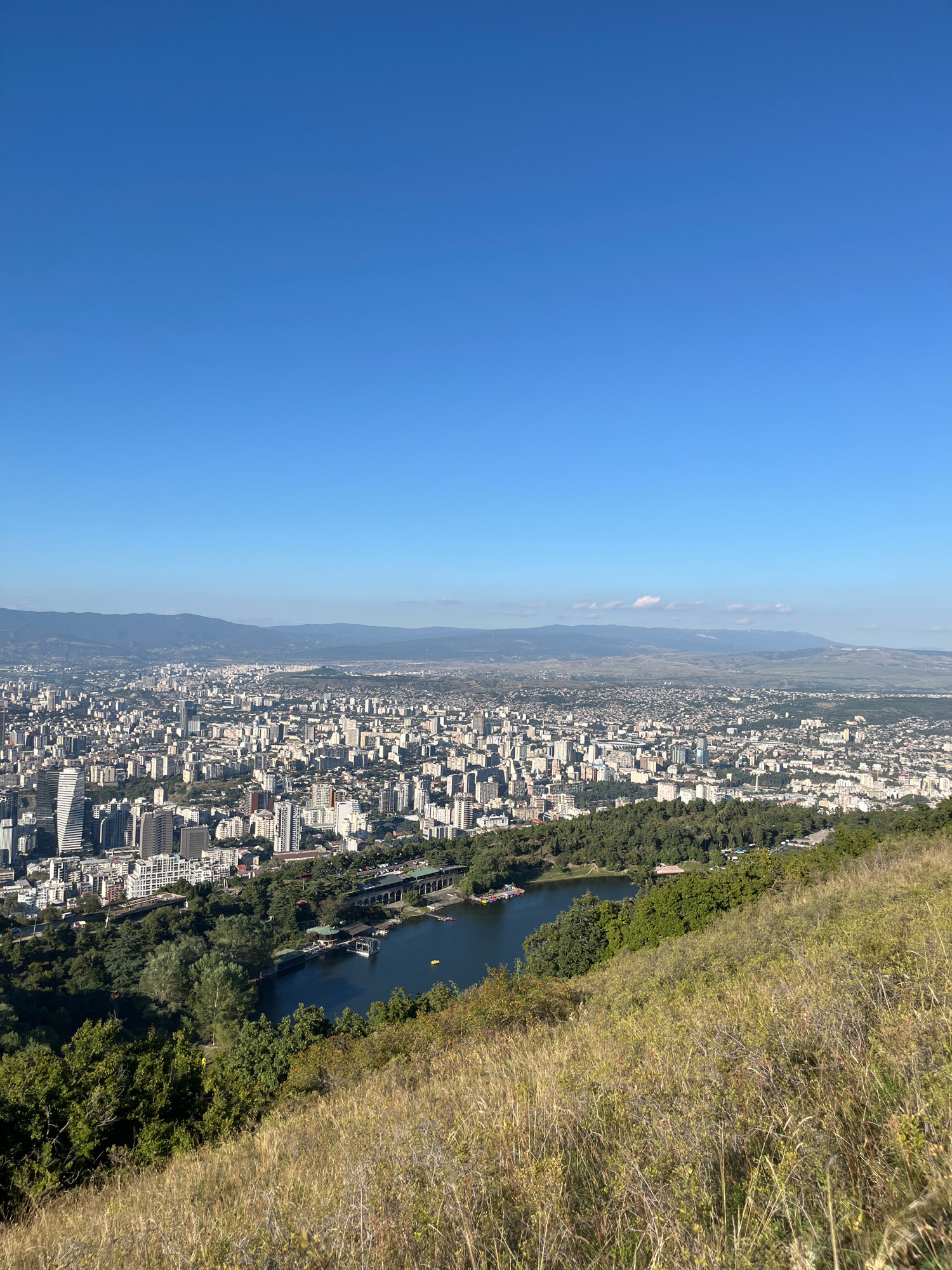 The image captures a panoramic view of Tbilisi, Georgia, from a high vantage point overlooking the city. The foreground is dominated by a grassy hillside with dry, tall grass and sparse vegetation. Beyond the hillside, a sprawling urban landscape unfolds, characterized by a dense collection of mid-rise and high-rise buildings in various shades of white and gray. The city stretches towards the horizon, where a range of hazy, undulating mountains form the backdrop under a clear, bright blue sky.
In the mid-ground, a prominent body of water, likely a lake or reservoir, is visible, fringed by lush green trees. Several structures, including what appears to be a boathouse or marina with boats docked, are situated along the edge of the water. A bridge with arches crosses a section of the water, and a road with a few vehicles is also discernible near the water's edge. The urban development extends beyond the greenery, with more buildings and infrastructure visible. The overall scene depicts a sunny day with good visibility, offering a sweeping vista of the city and its natural surroundings. No people are clearly identifiable in the image.
