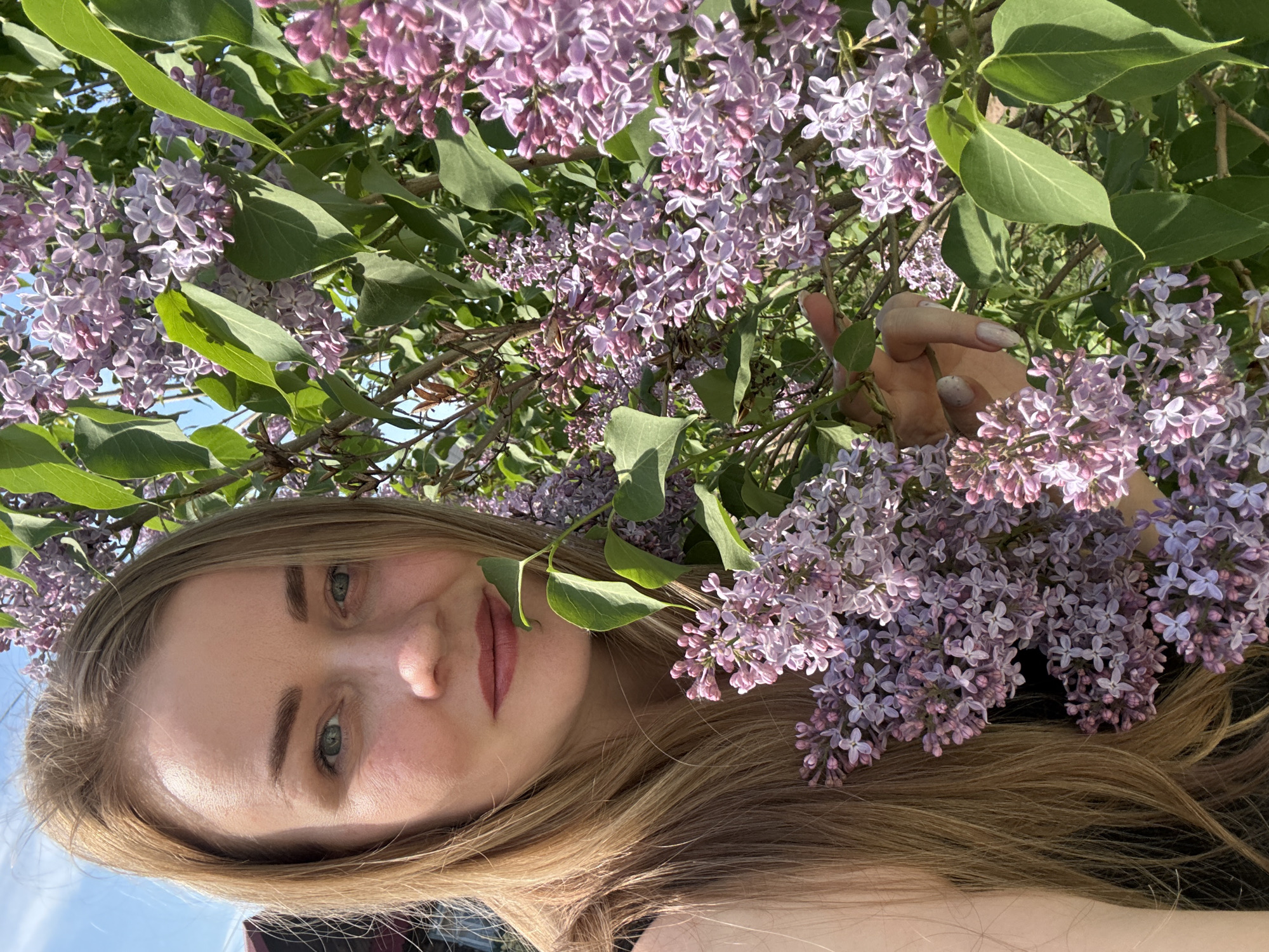 A young woman with long, light-brown hair is surrounded by lilac flowers and green leaves. She is looking at the camera with a slight smile. Her right hand is visible, holding one of the lilac branches. The sky is visible in the background. The location is Chaikovsky, Russia.