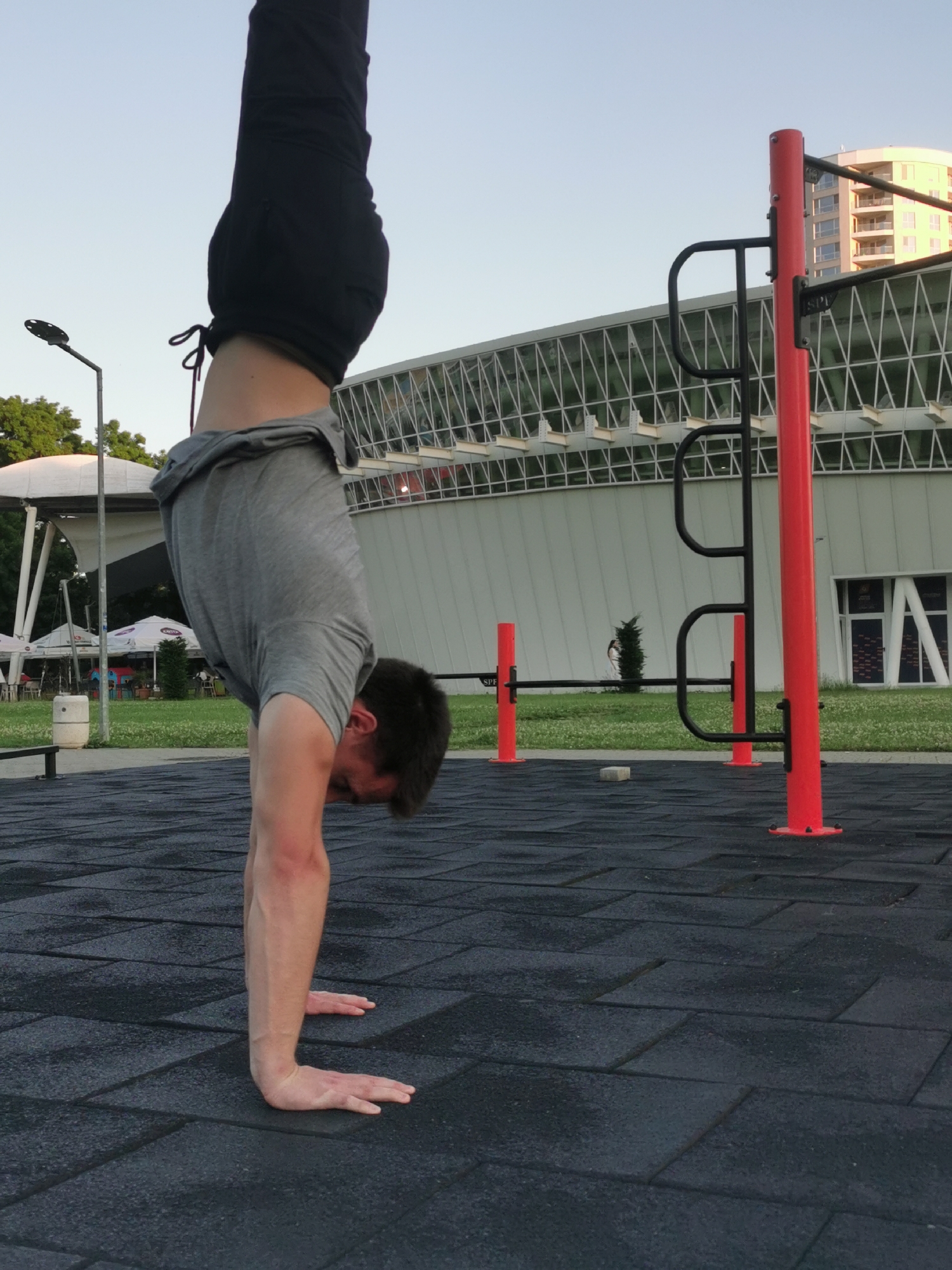 A man is performing a handstand in a park. He is wearing black shorts and a gray t-shirt. The park has black rubber tiles. In the background, there is a modern building with a white facade and triangular window design. There is also a red metal structure with black handles, typical of outdoor fitness equipment. Other elements include a street lamp, trees, and a building with a white tent in front of it, possibly a cafe. In the far background, a taller building is visible. Based on the architecture and park amenities, this scene is likely set in or near Sea Garden Park in Burgas, Bulgaria.