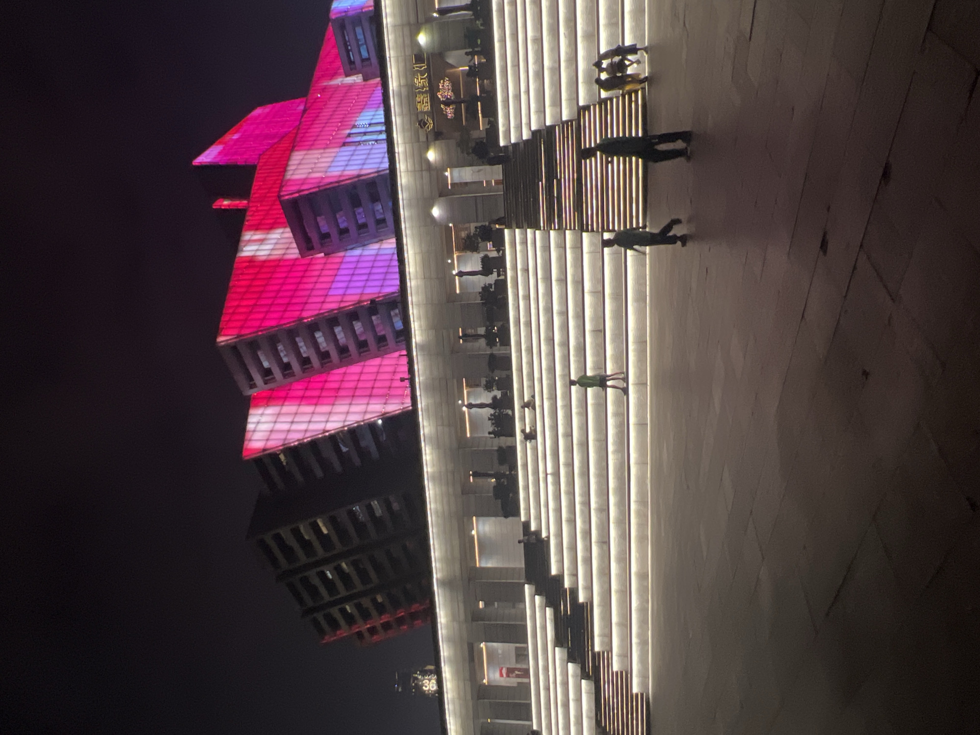 An exterior, nighttime shot of a modern building complex in Jiangbeicheng Subdistrict, China. The building's facade is illuminated with vibrant pink and white lights, creating a striking geometric pattern. In front of the building, a wide, multi-tiered staircase is lit with soft, white lights, with several people scattered across its steps and the plaza below. To the right of the stairs, a group of three people are visible, one of whom appears to be interacting with a child. The plaza itself is paved with large, dark stone tiles. The overall scene suggests a public space designed for gathering and entertainment.